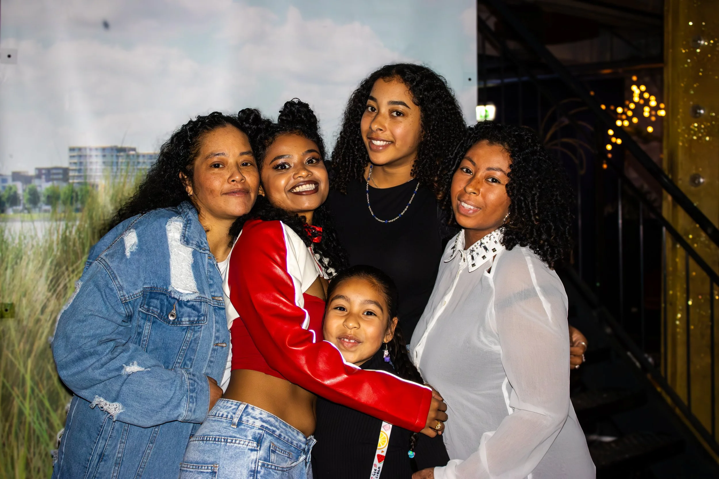 Group of five women and one girl smiling and hugging together at an indoor event.