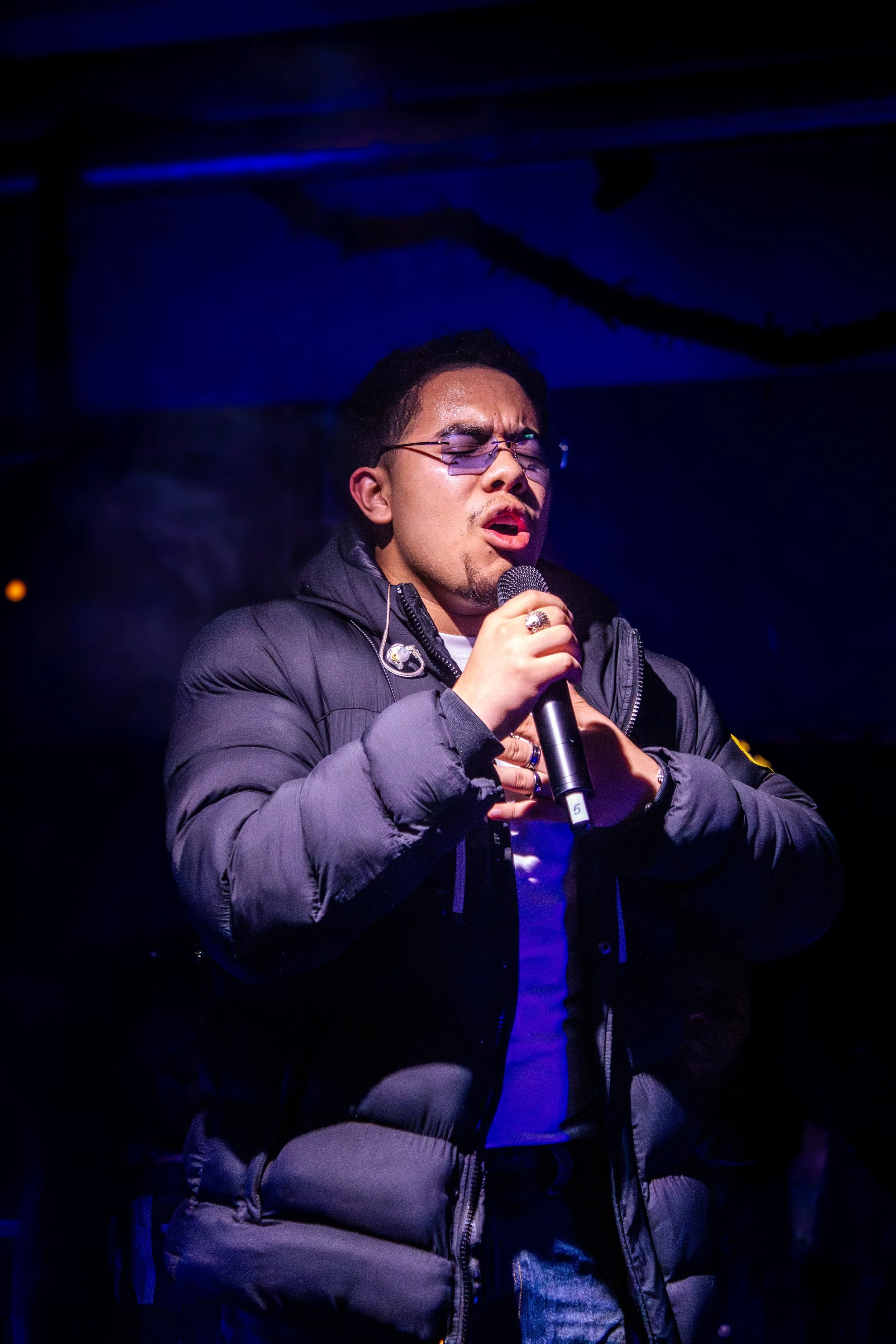 A man singing into a microphone at a dark indoor venue, wearing sunglasses and a puffy black jacket.