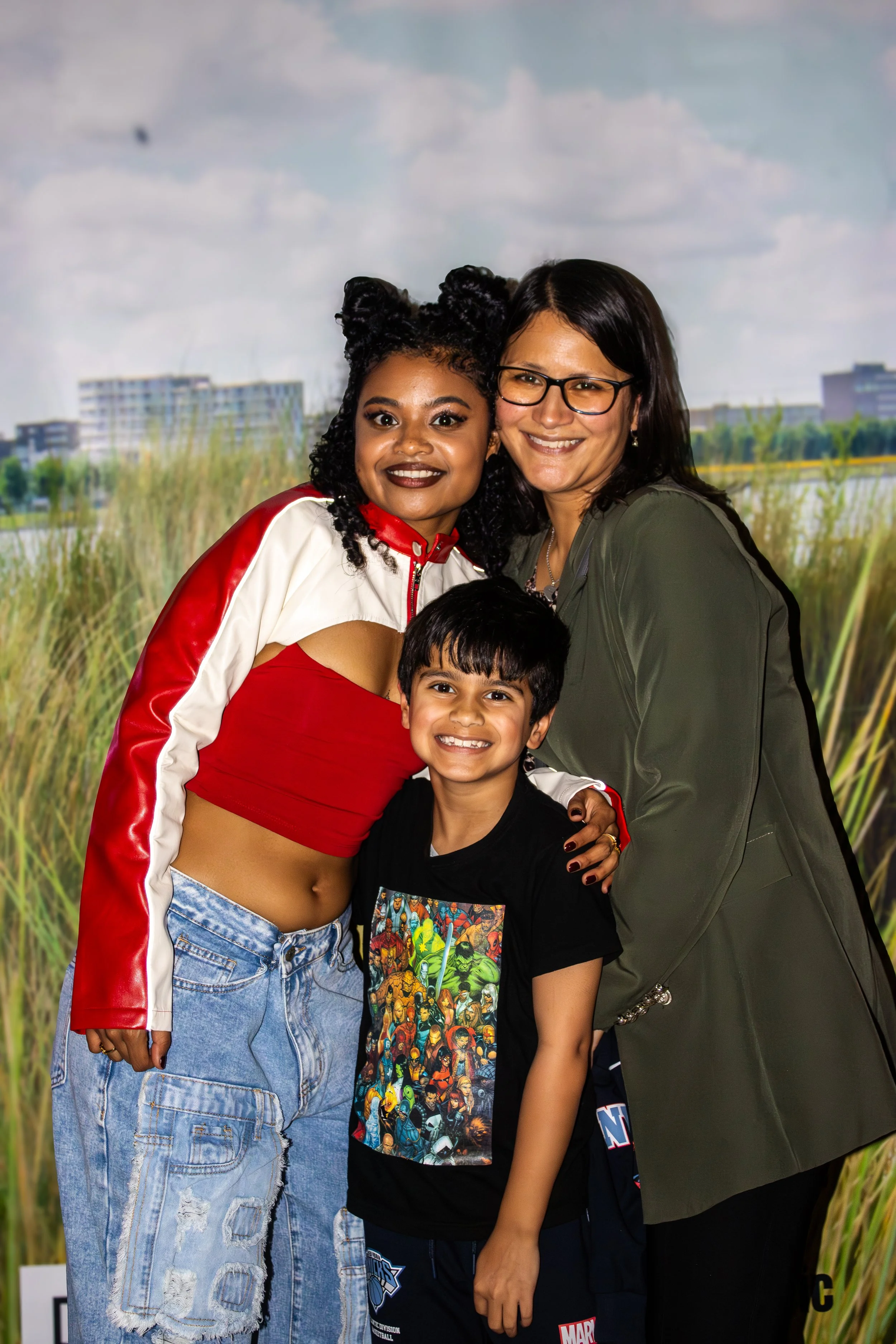 Three people: a young woman with curly hair in a red and white jacket and denim pants, a smiling woman wearing glasses and a green jacket, and a smiling young boy in a Marvel T-shirt, standing outdoors with tall grass and buildings in the background.