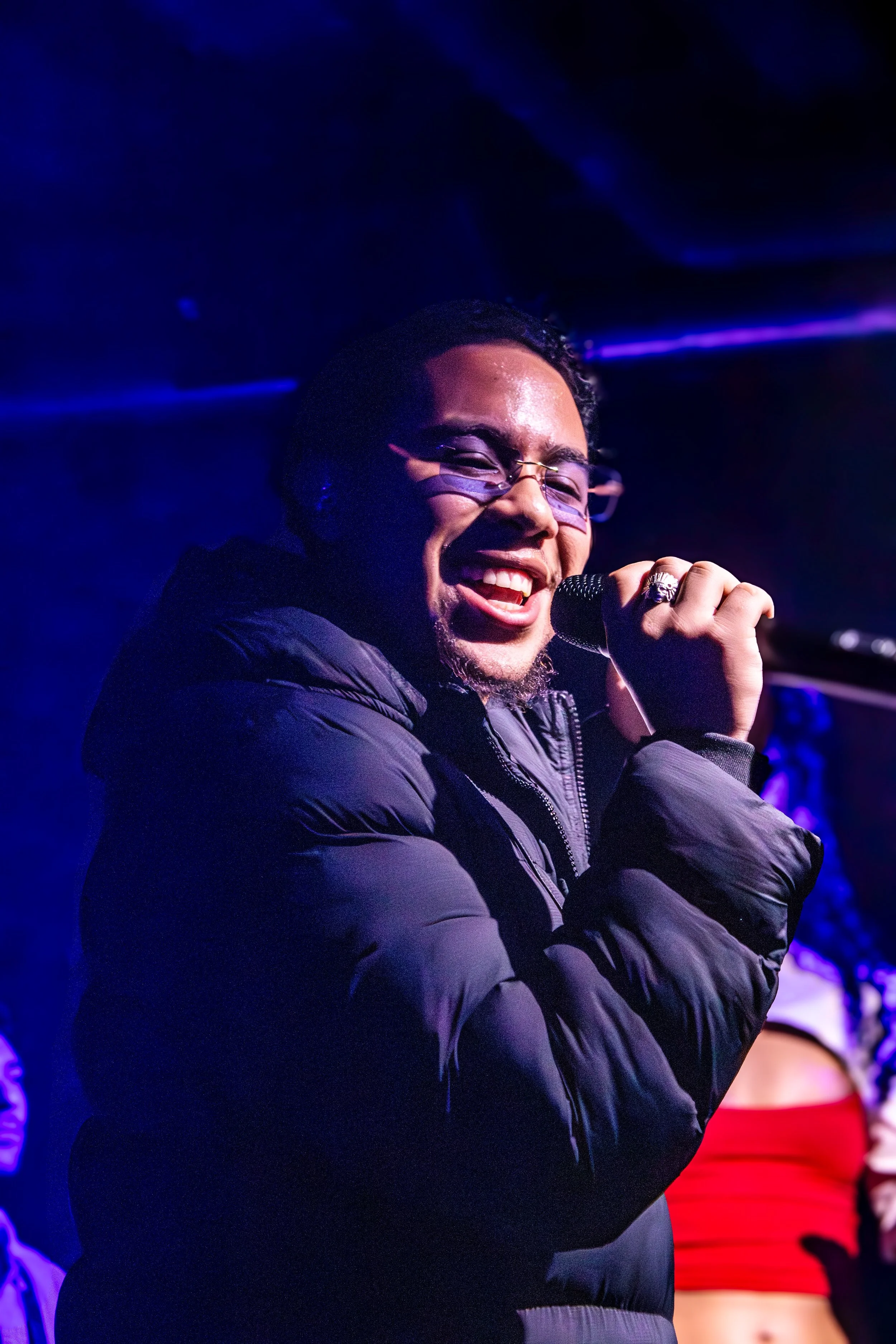 A man smiling and singing into a microphone on stage, wearing glasses and a black puffer jacket, with purple and blue stage lighting.