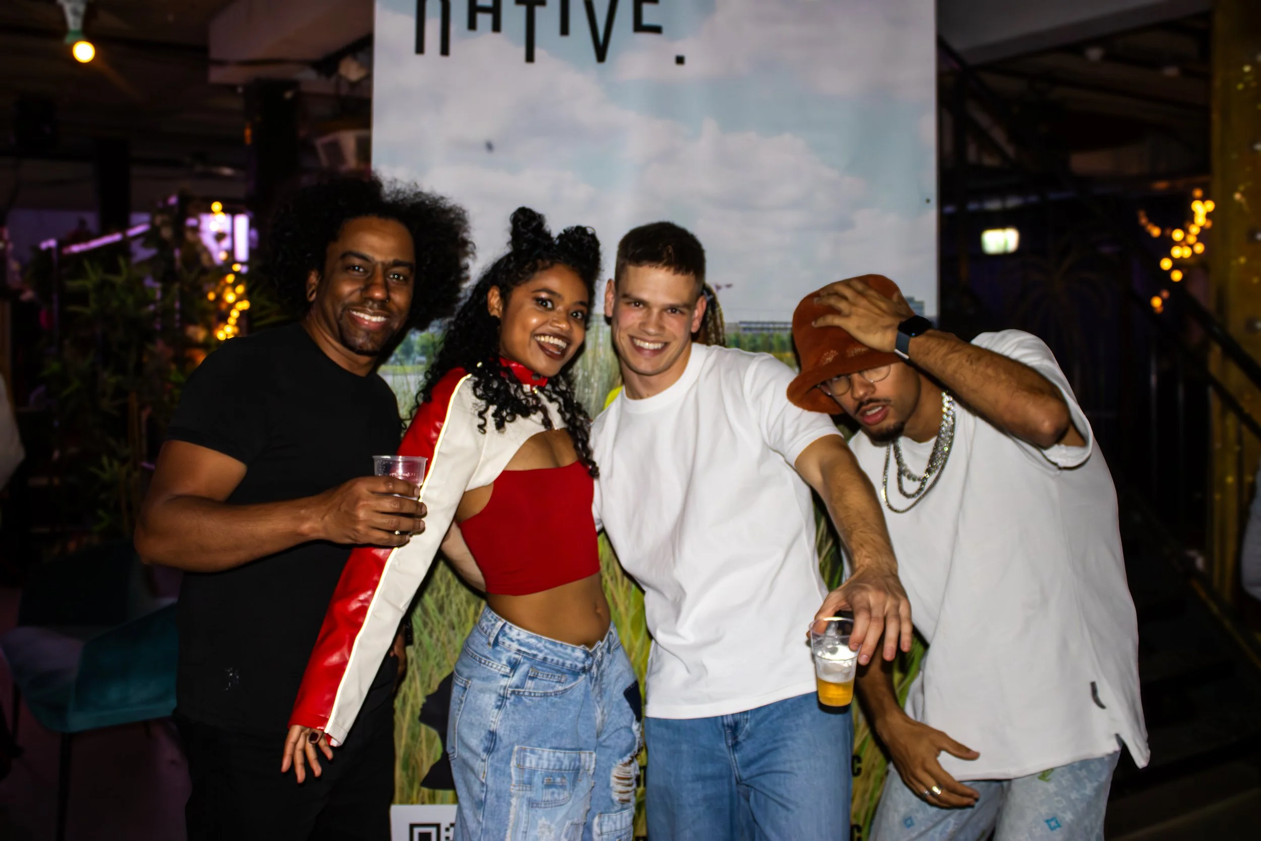 Four people standing together at a party, smiling and holding drinks, with a large banner in the background.