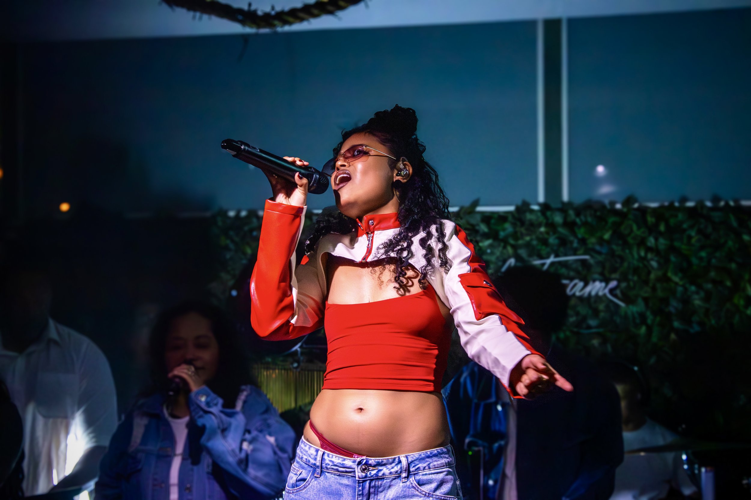 A woman singing into a microphone on stage, wearing a red and white striped jacket with a red crop top, red sunglasses, and denim shorts, with a green leafy backdrop and other people in the background.