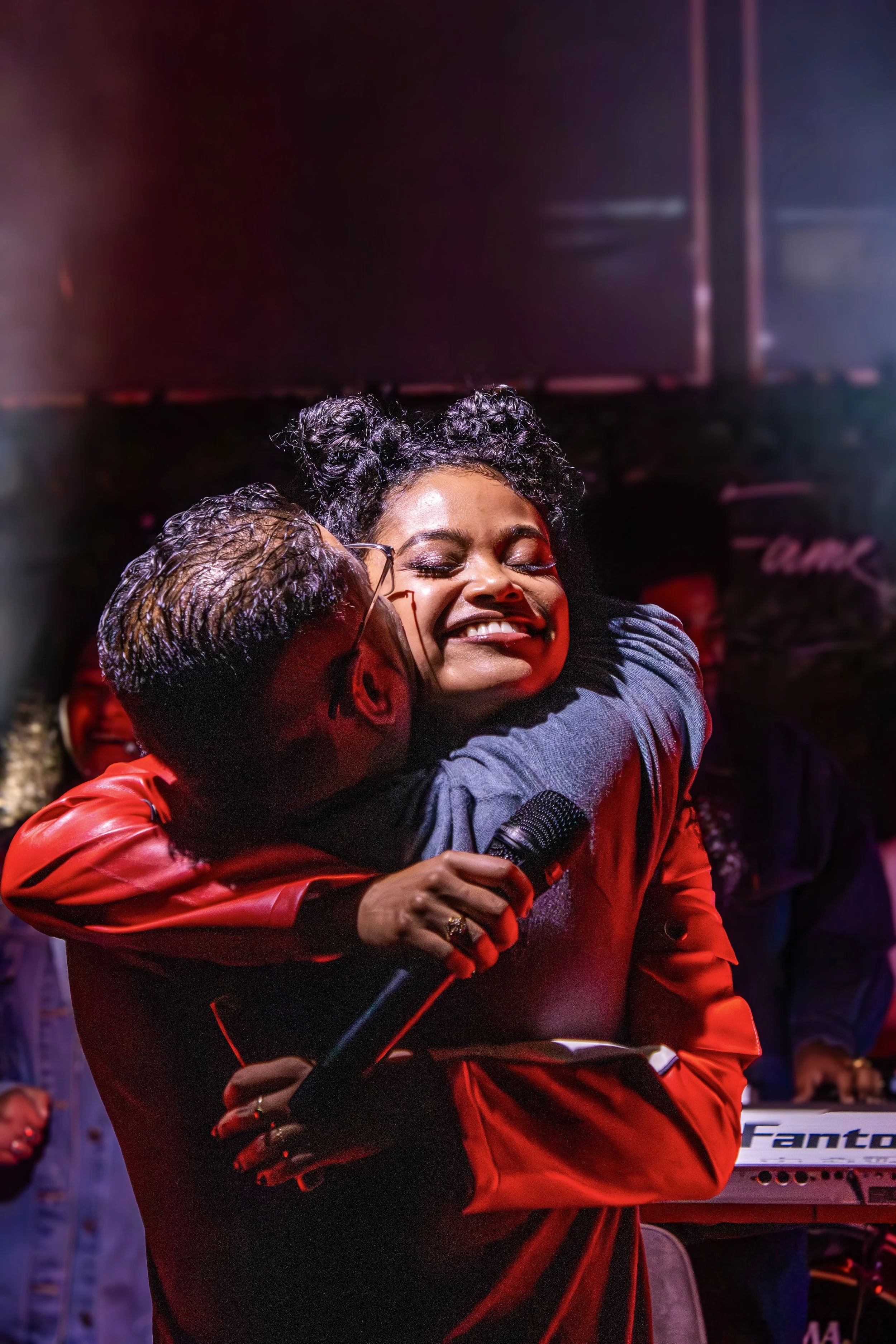 A woman with curly hair and closed eyes hugging a man while holding a microphone, smiling, on stage with musical equipment in the background.