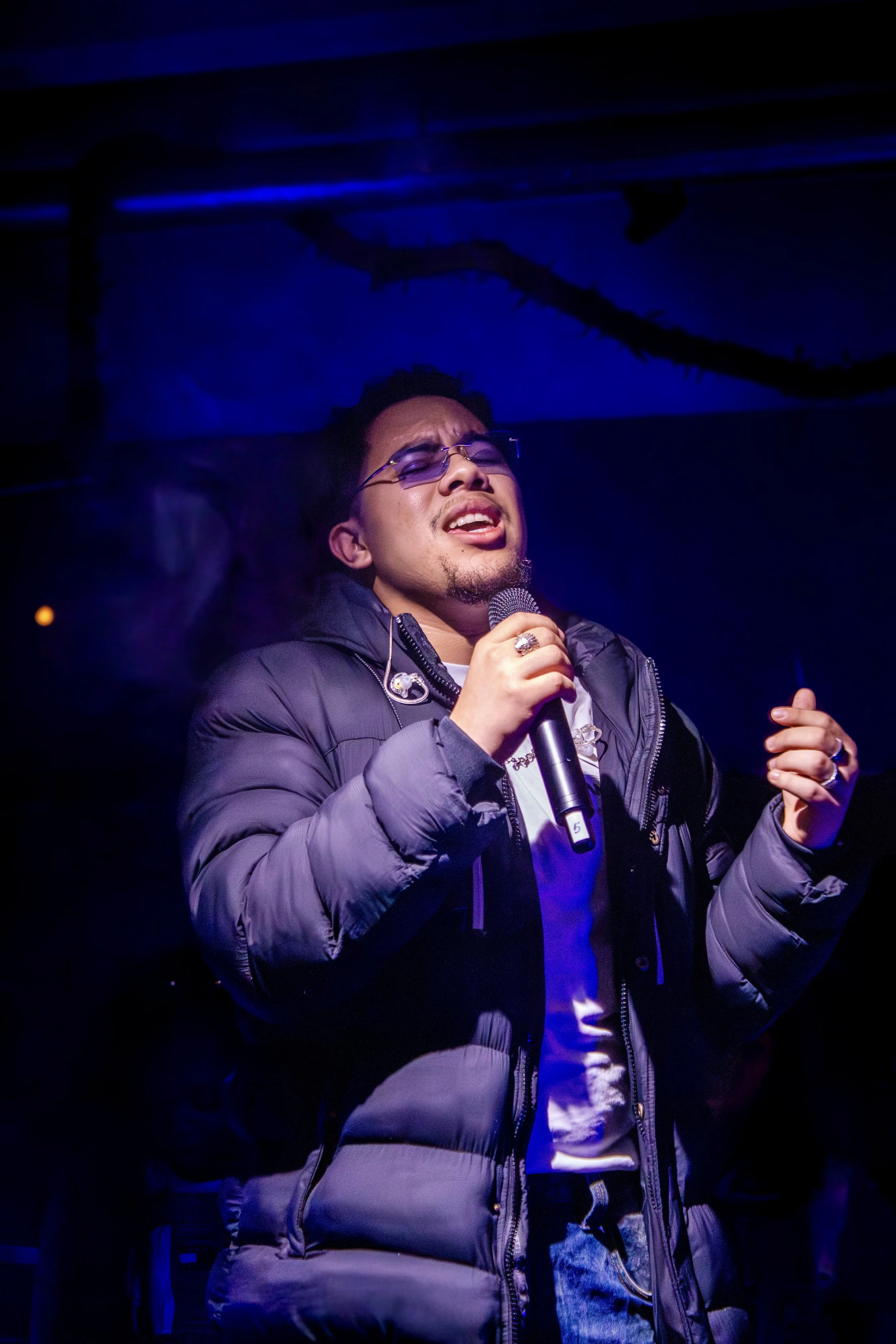 A man with glasses singing passionately into a microphone on stage, wearing a dark padded jacket and a white shirt, with a dark background and blue lighting.
