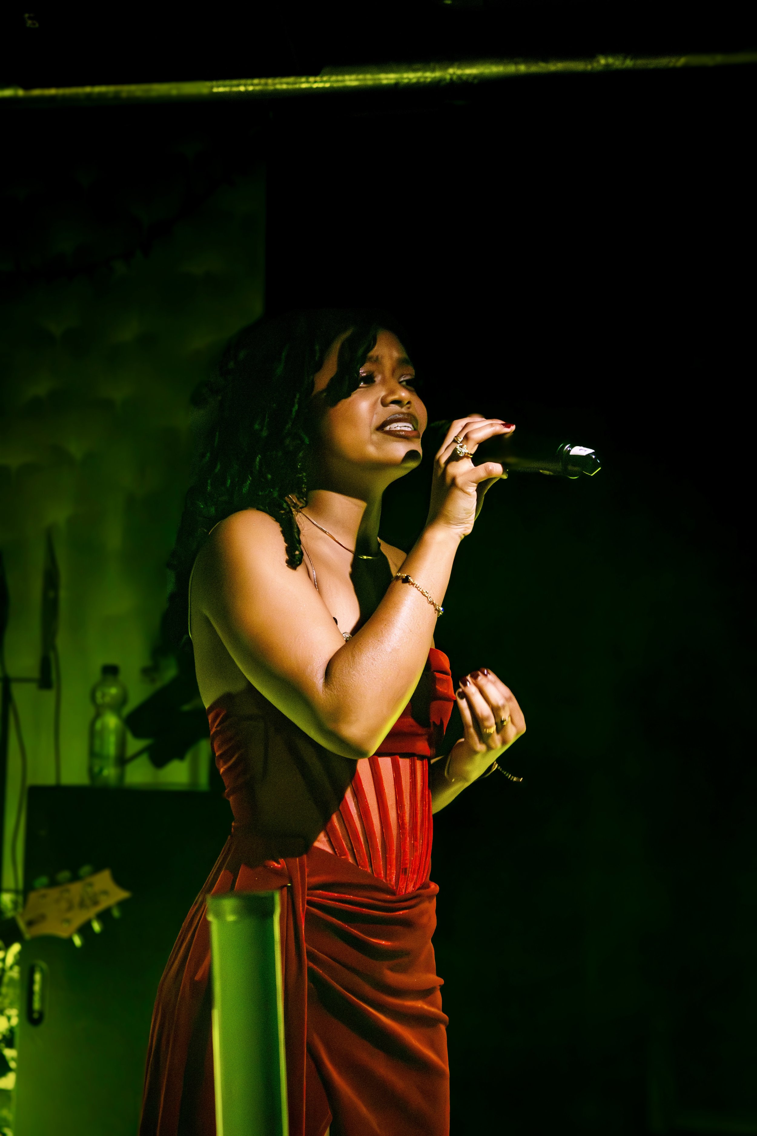 A woman with dark curly hair singing into a microphone on stage, dressed in a red and black strapless dress, with stage lighting highlighting her.