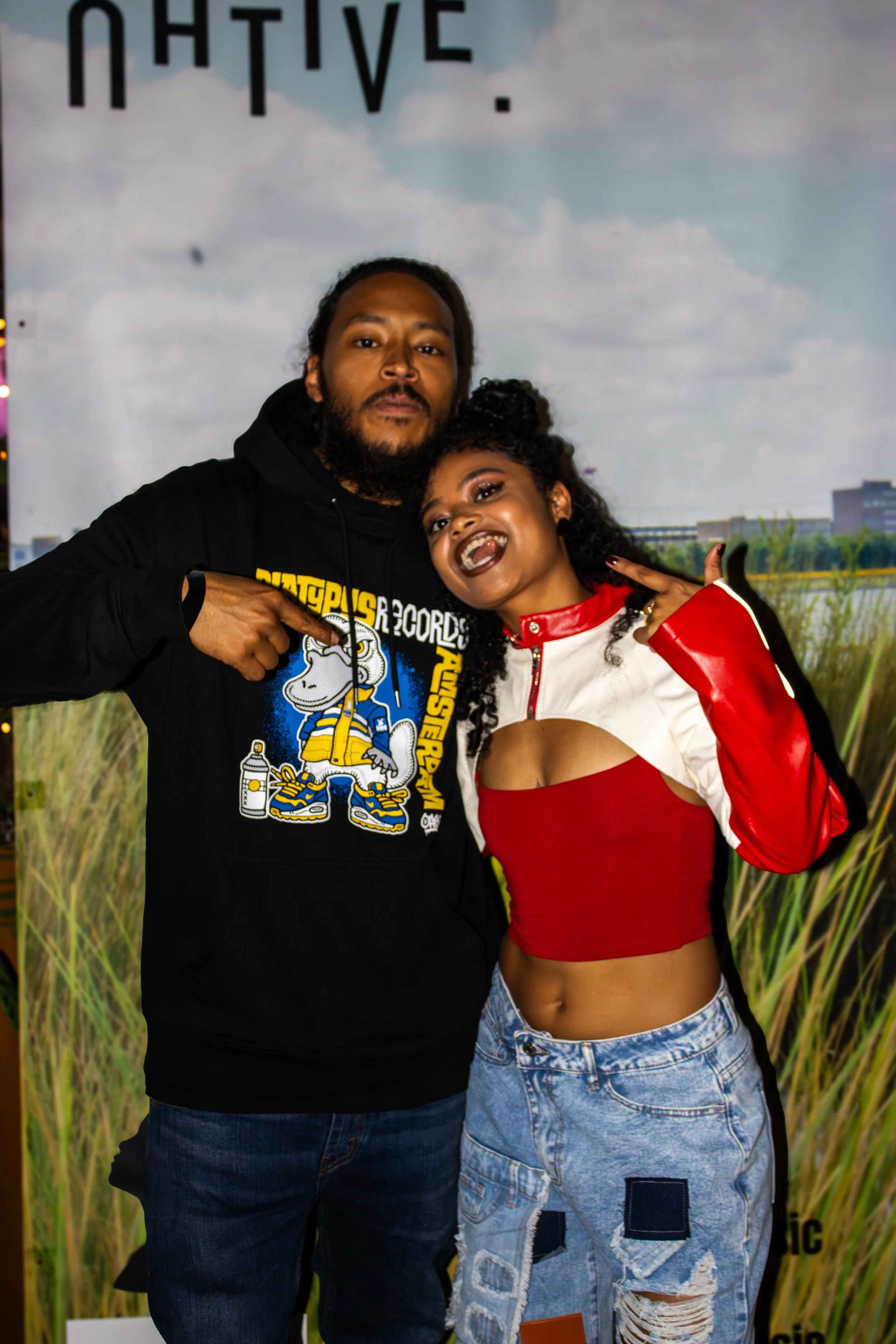 A man and woman posing together, with the man pointing at the woman who is smiling through her teeth and wearing a red and white jacket with ripped jeans. The background features a natural scene with grass and a cloudy sky with a city skyline in the 