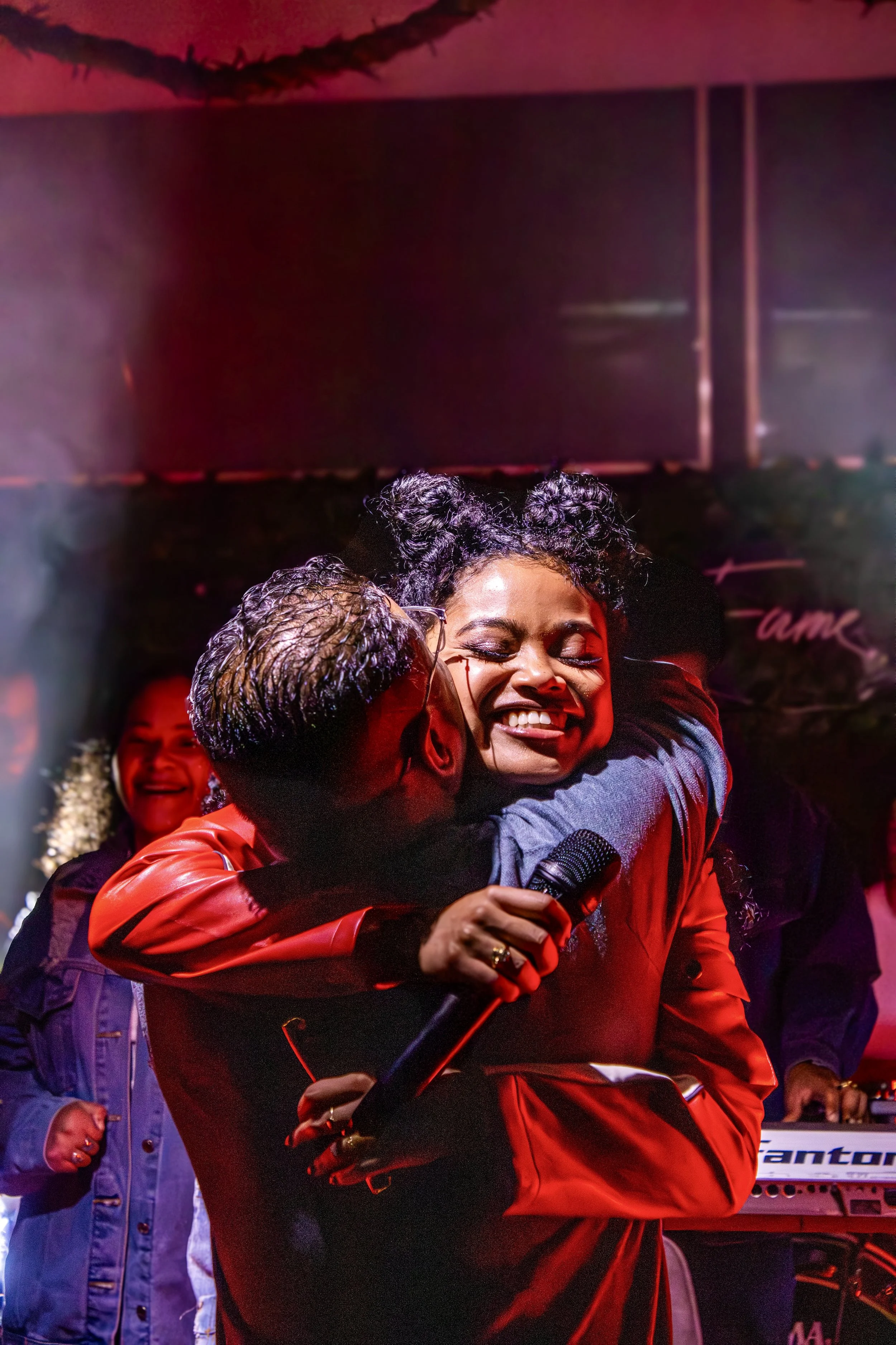 A woman with curly hair and a black top hugging a man in a red jacket on stage, smiling with a microphone in her hand, amid others smiling in the background, during a lively event with colorful lighting.