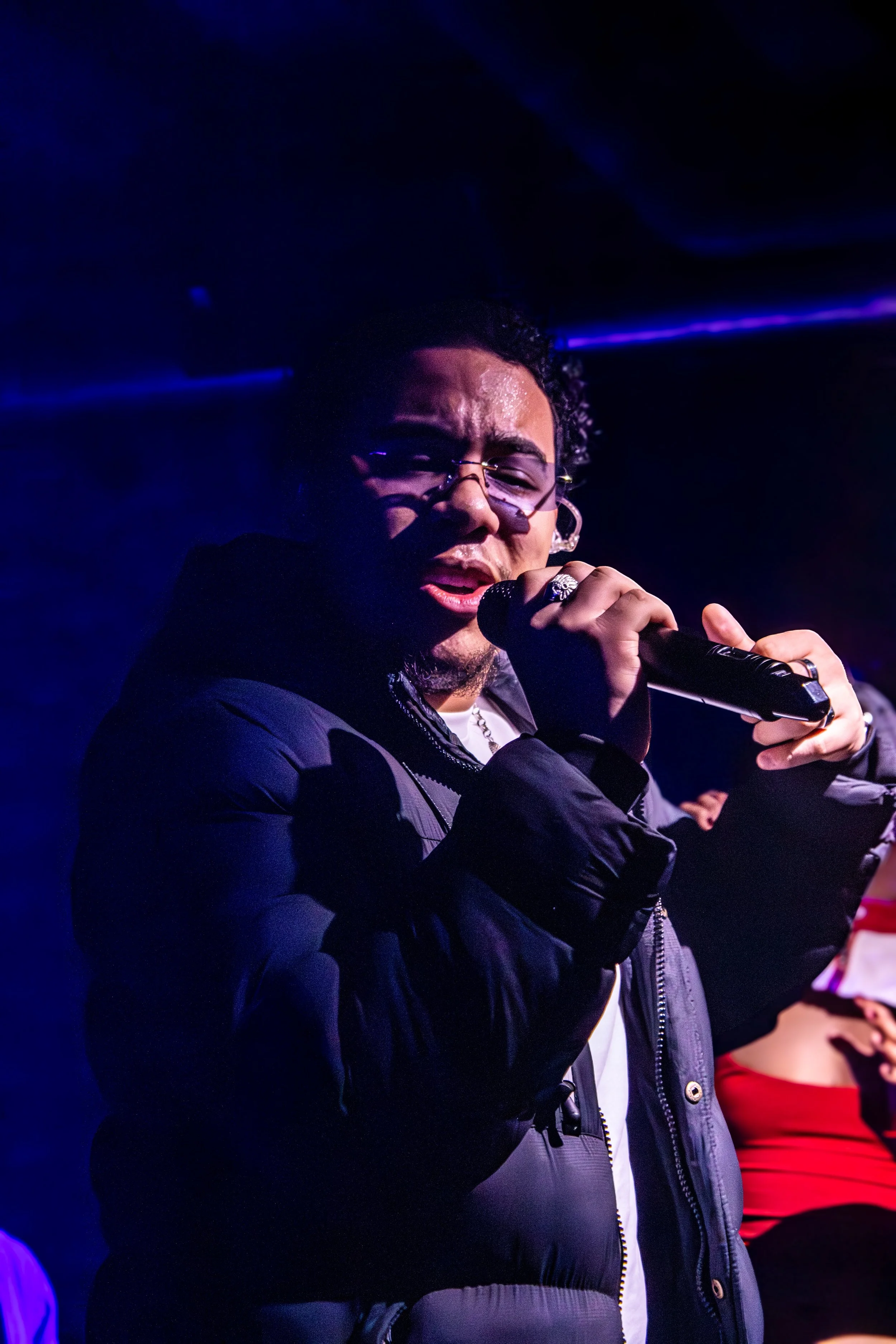 A male singer wearing glasses and a black jacket performing with a microphone on stage, with blue and purple lighting.