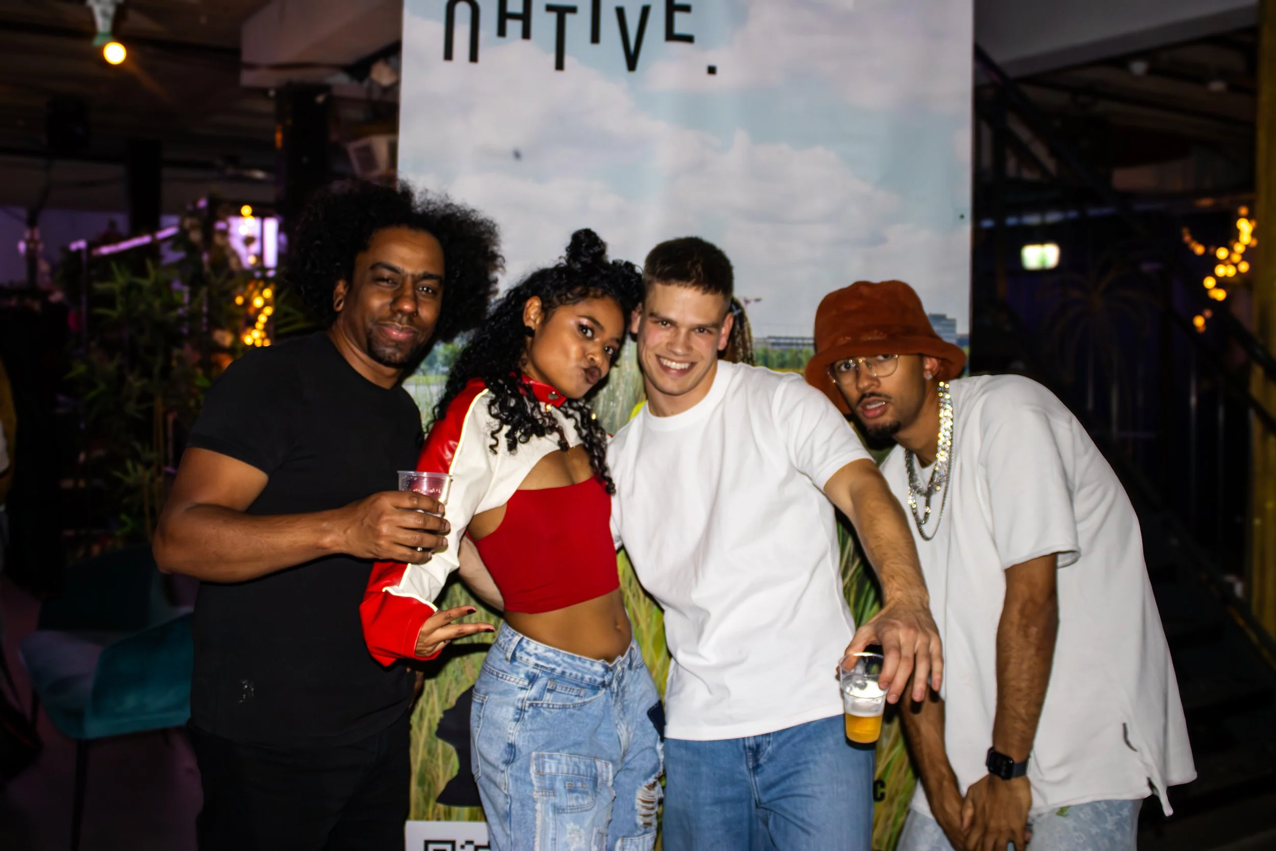 Four people at a social event standing close together, smiling, and holding drinks. They are indoors with a background banner that partially reads 'NATIVE' and decorative lighting.