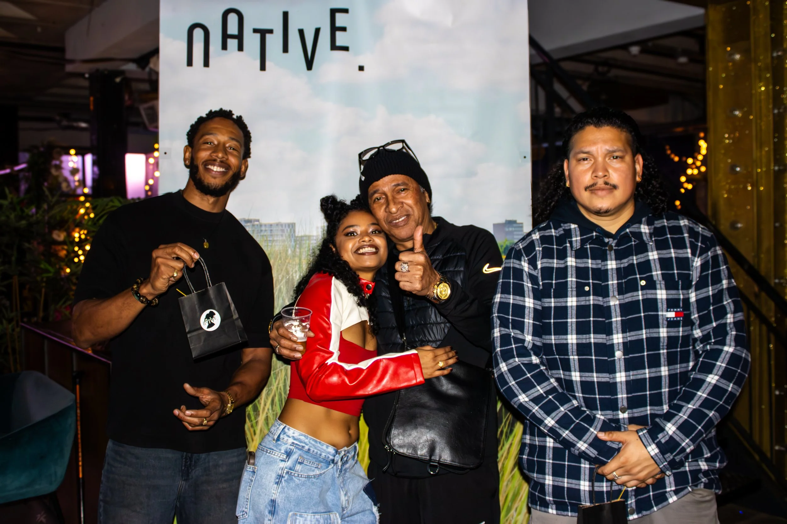 Four people standing together at an event, smiling and posing for the camera, with a large banner behind them that says 'NATIVE.' The group includes two men and one woman in the middle, all smiling, and a man on the right with a neutral expression. T
