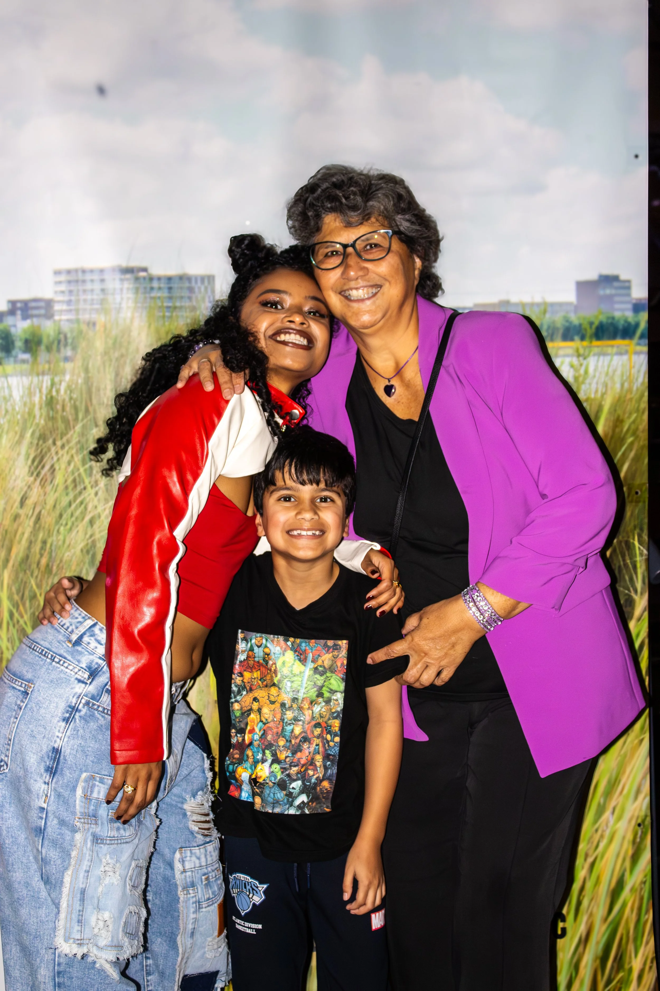 Three people, an older woman and two children, hugging and smiling in front of a nature-themed backdrop with grass and buildings in the background.