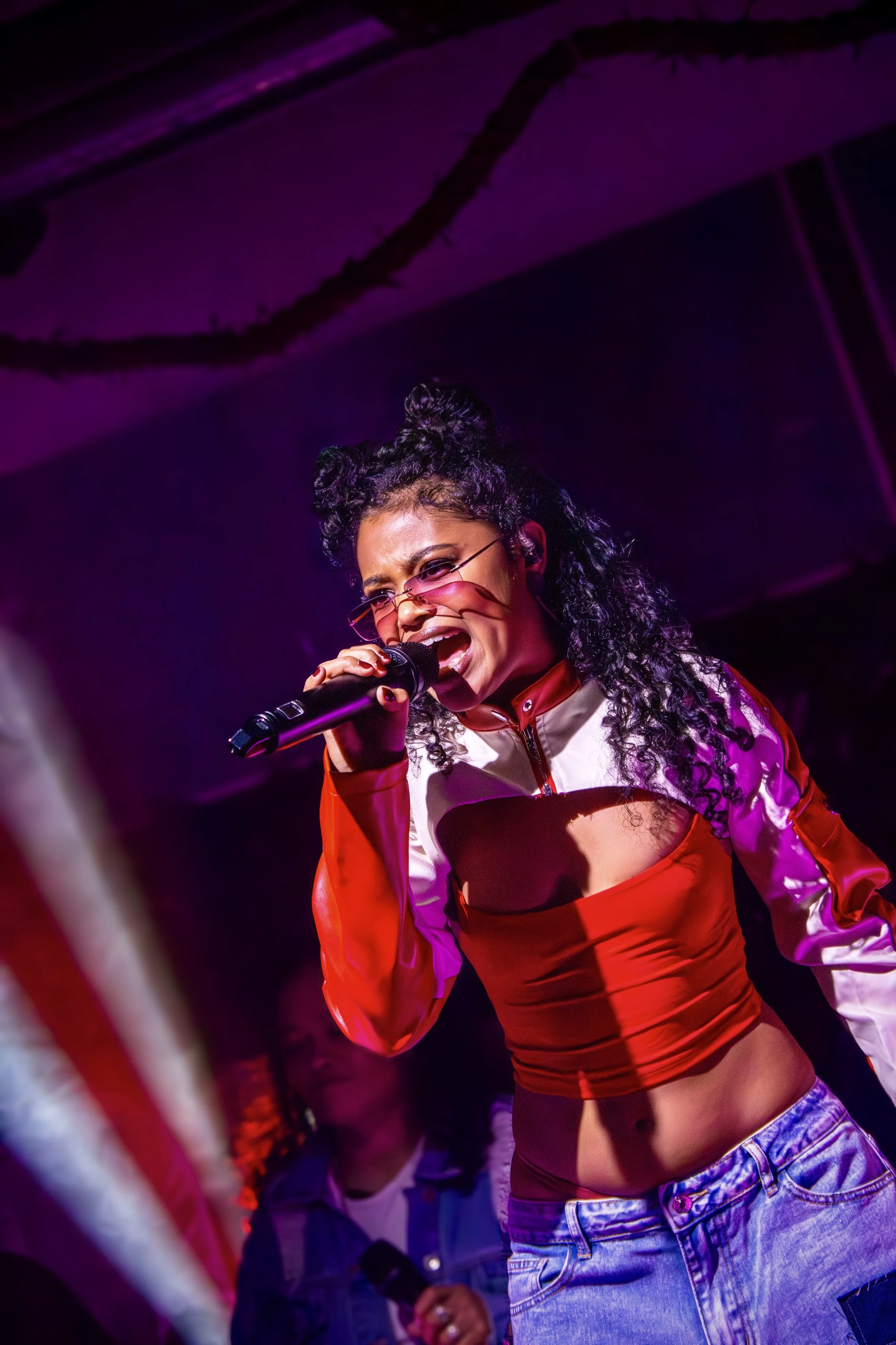 A woman singing into a microphone on stage under purple lighting, wearing pink tinted sunglasses, a red and white jacket, and blue jeans, with curly hair styled in two buns.