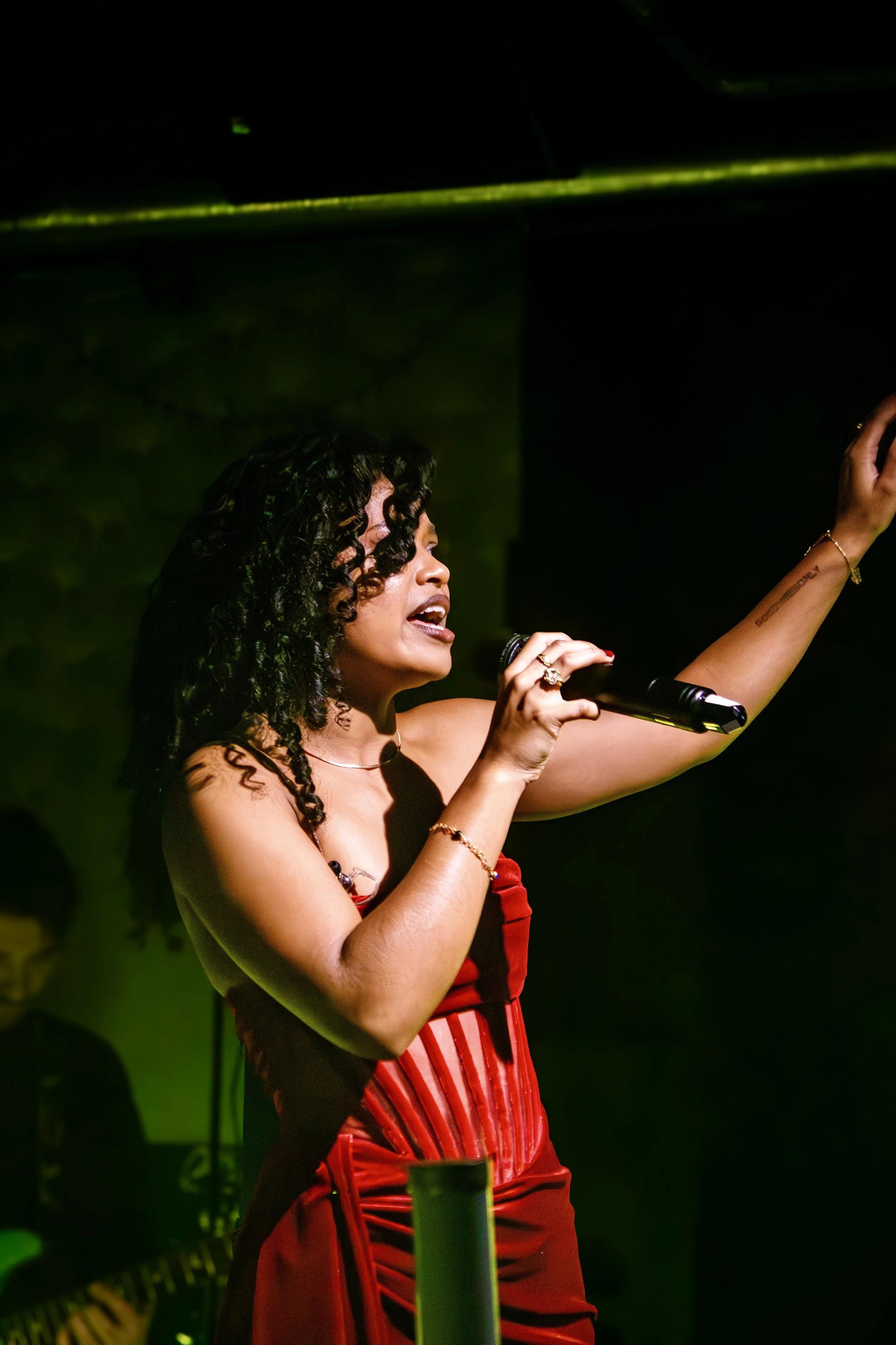 A woman singing passionately into a microphone on stage, wearing a red dress, with curly black hair, in a dark setting with green and yellow lighting.