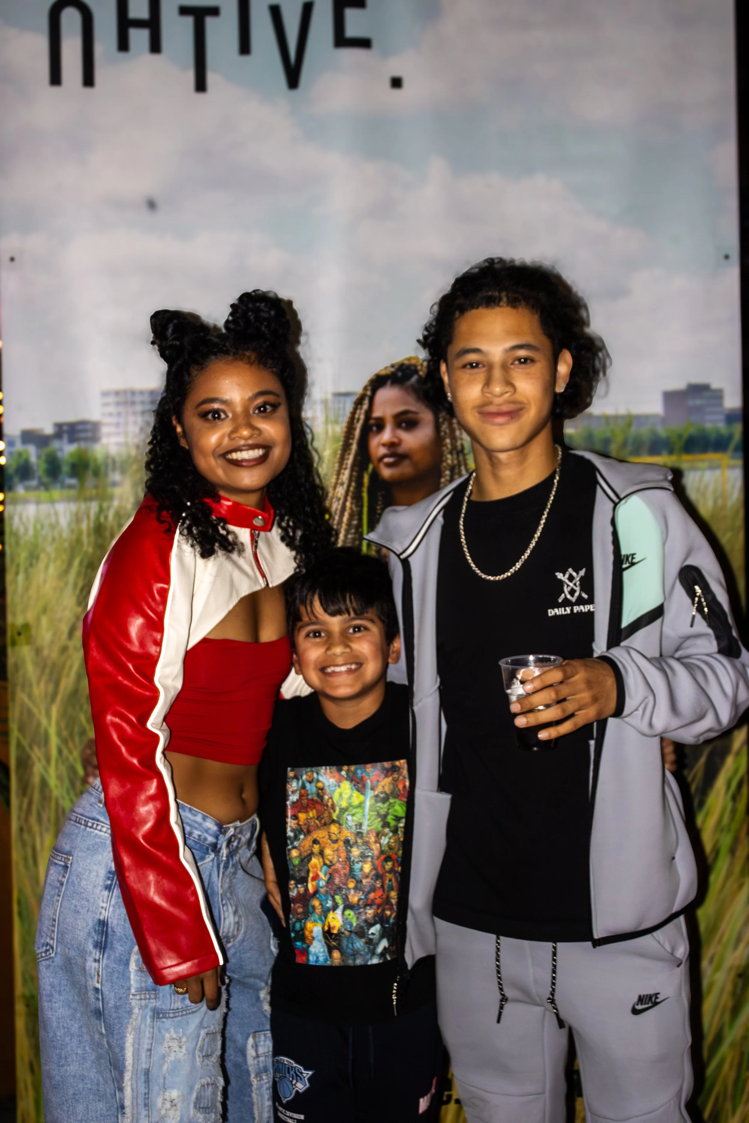Group of four diverse people, two women and two boys, standing together outdoors during daytime with a backdrop of a field and city buildings, smiling at the camera.