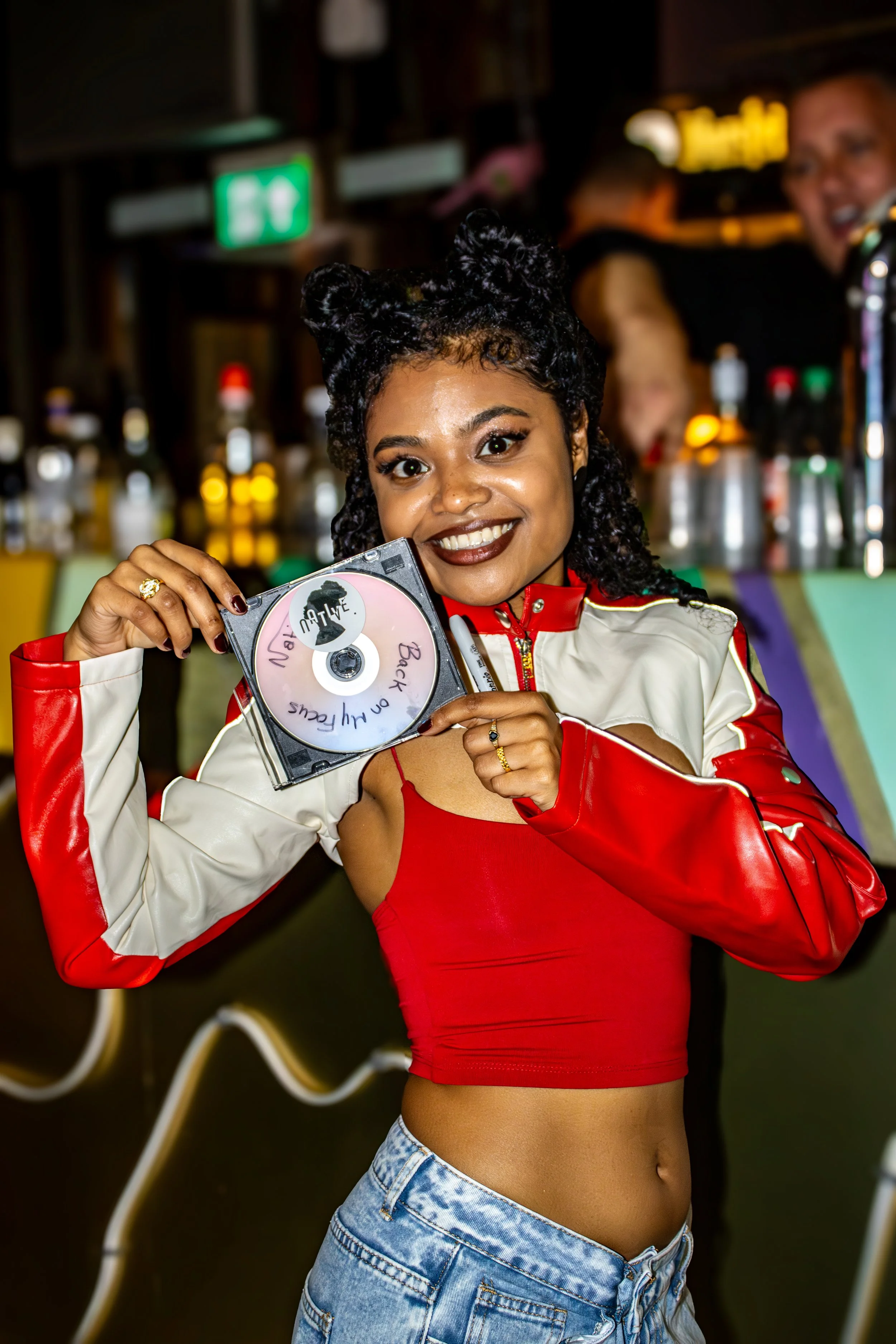 A young woman with curly black hair and dark lipstick is smiling and holding a CD case labeled 'Back to the Future.' She is wearing a red tank top and a color-blocked jacket with red and white sleeves, and is in a bar or club setting with colorful li