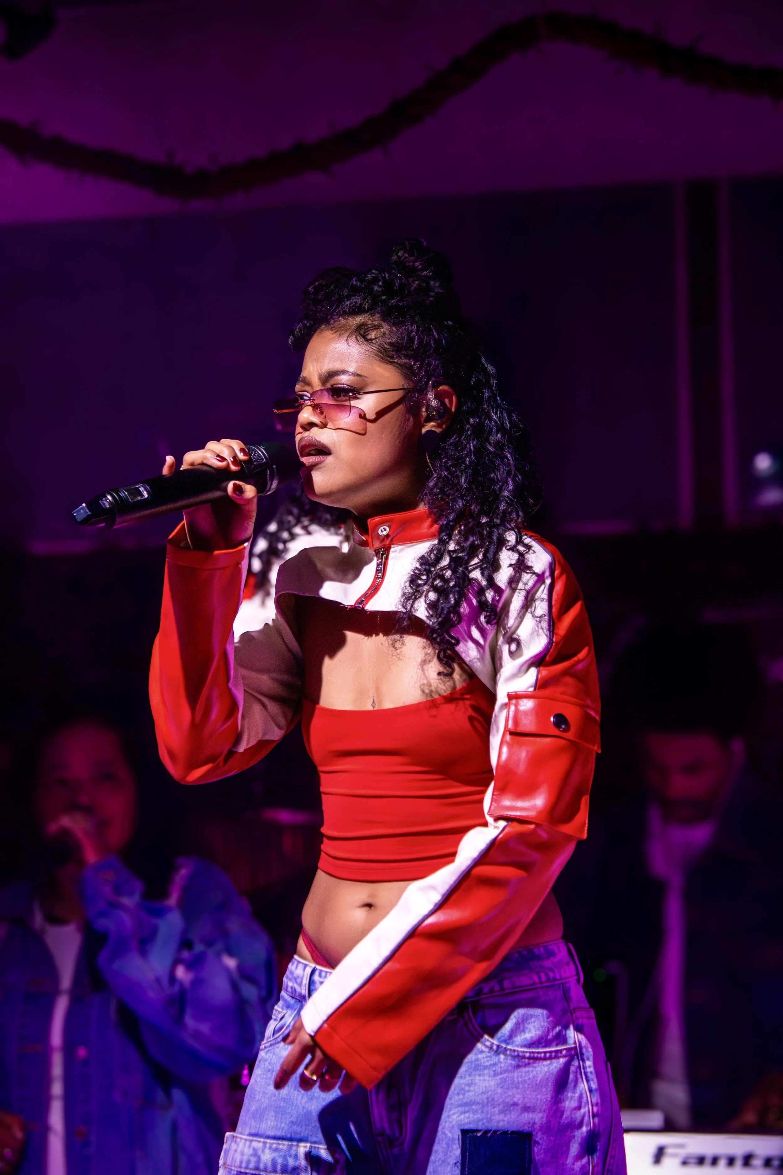 A woman with curly black hair and pink-tinted glasses performs on stage, holding a microphone, wearing a red and white cropped jacket and baggy jeans.