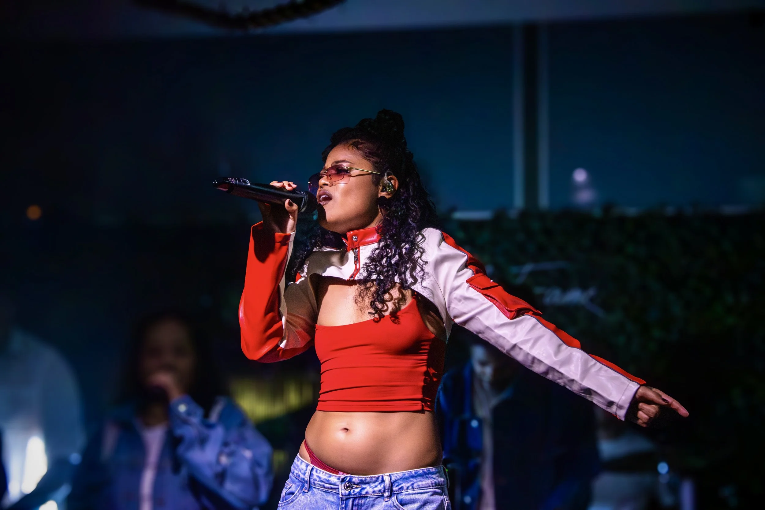 A female singer performing on stage, wearing a red crop top, a sporty jacket, and sunglasses, holding a microphone and singing passionately.
