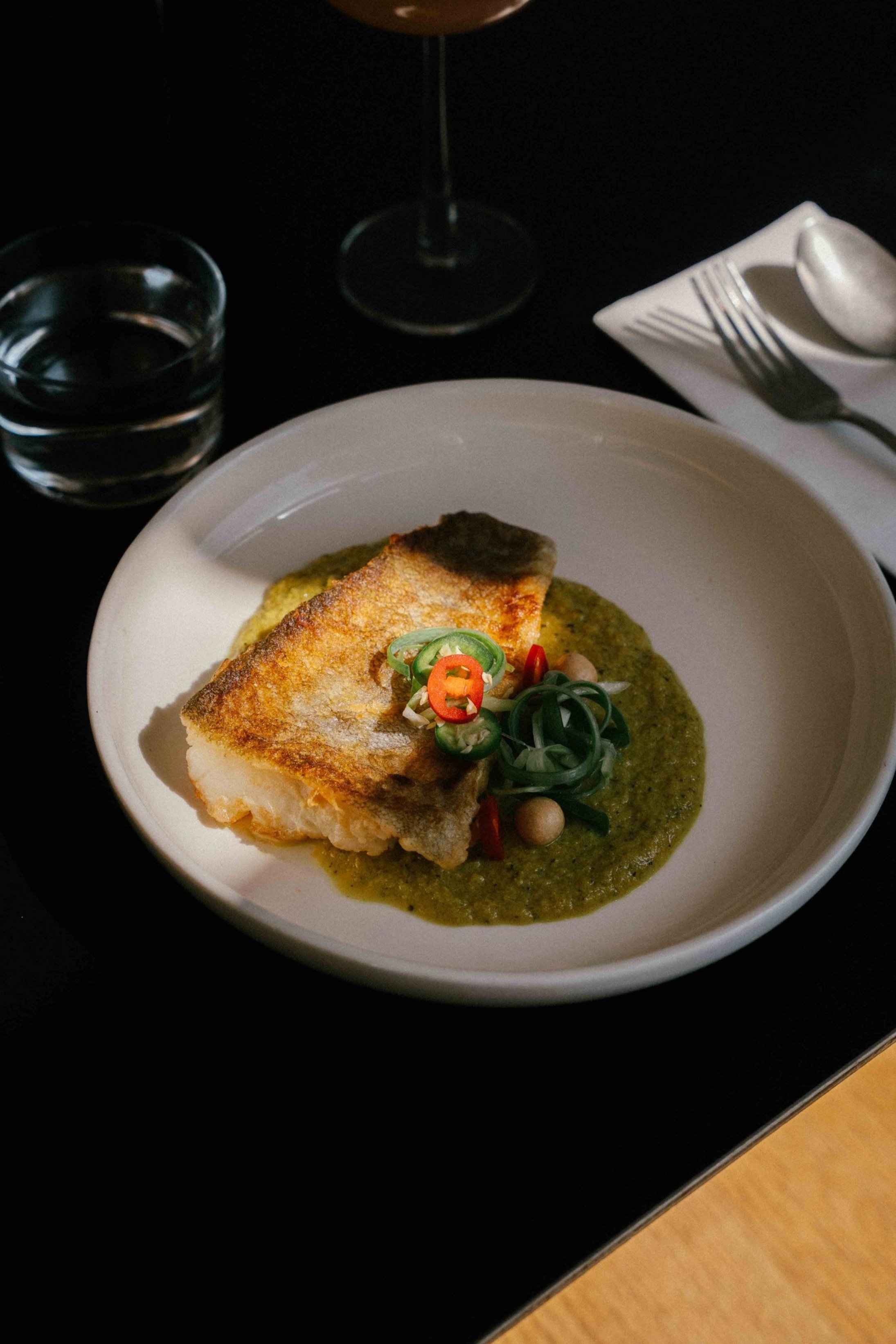 A plate of cooked fish fillet served over green sauce with sliced green and red chili peppers and small round vegetables, accompanied by a glass of red wine, a glass of water, and a fork and spoon on a white napkin.