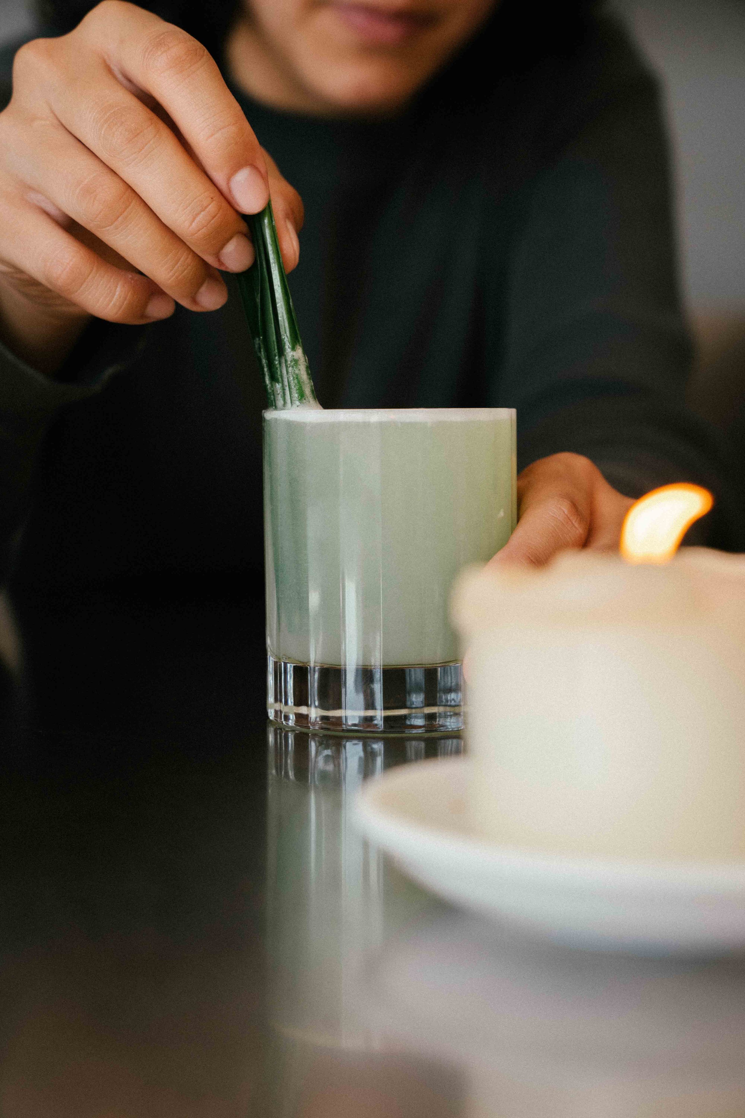 A person stirring a green beverage in a glass with a green straw, with a candle on a white plate in the foreground.