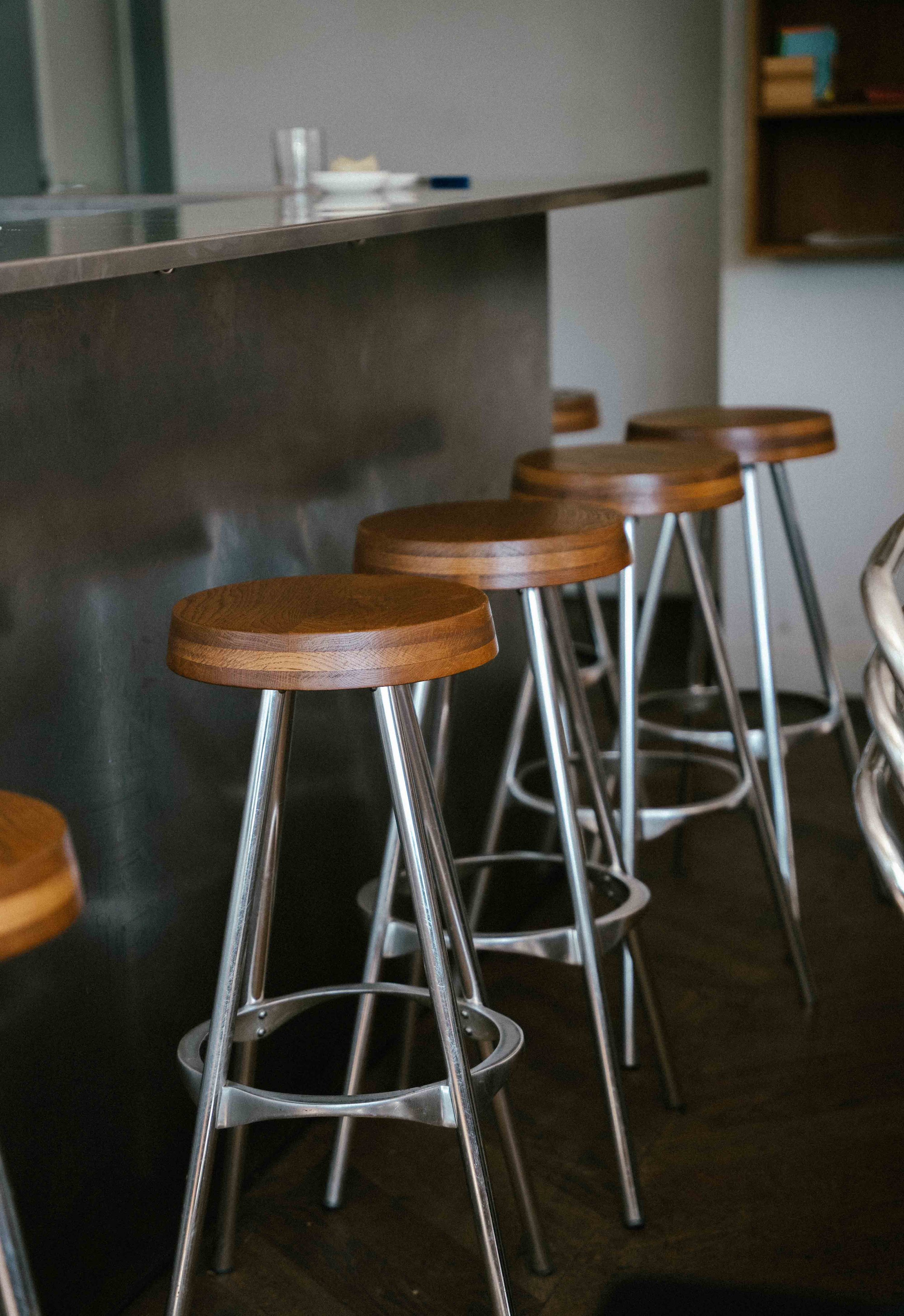 Four wooden and metal bar stools lined up along a dark counter in a cafe or bar, with a blurred background showing a glass and a small plate.