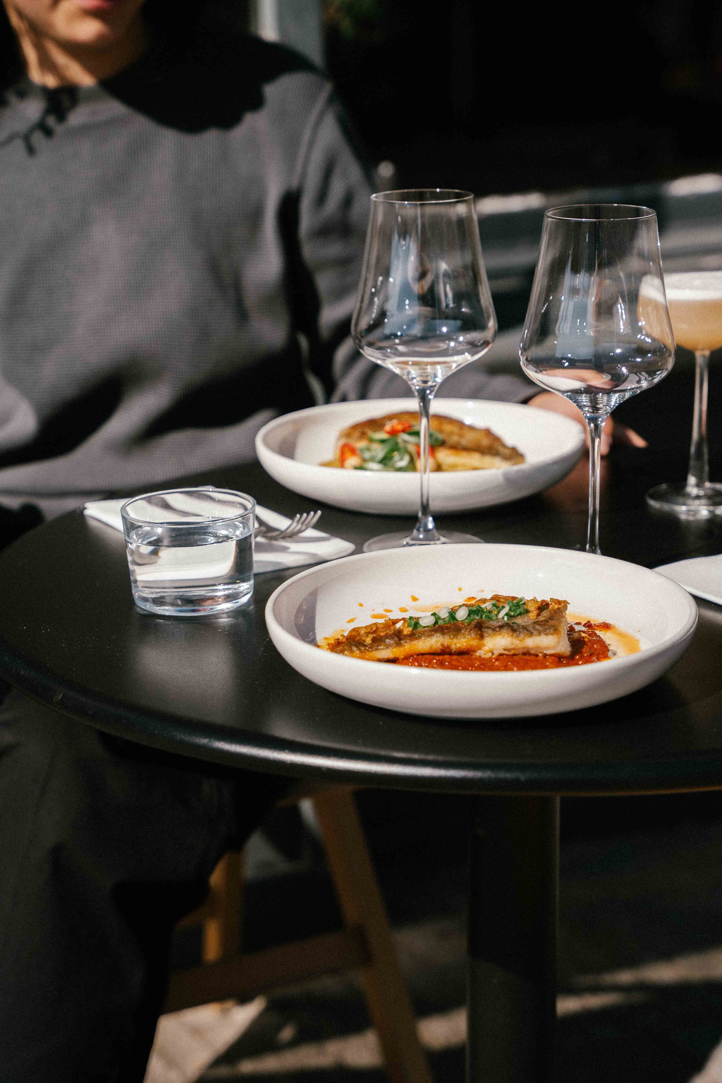 A black round table with two plates of food, two wine glasses, a glass of water, and a dessert cocktail, with a person seated nearby wearing a gray shirt.