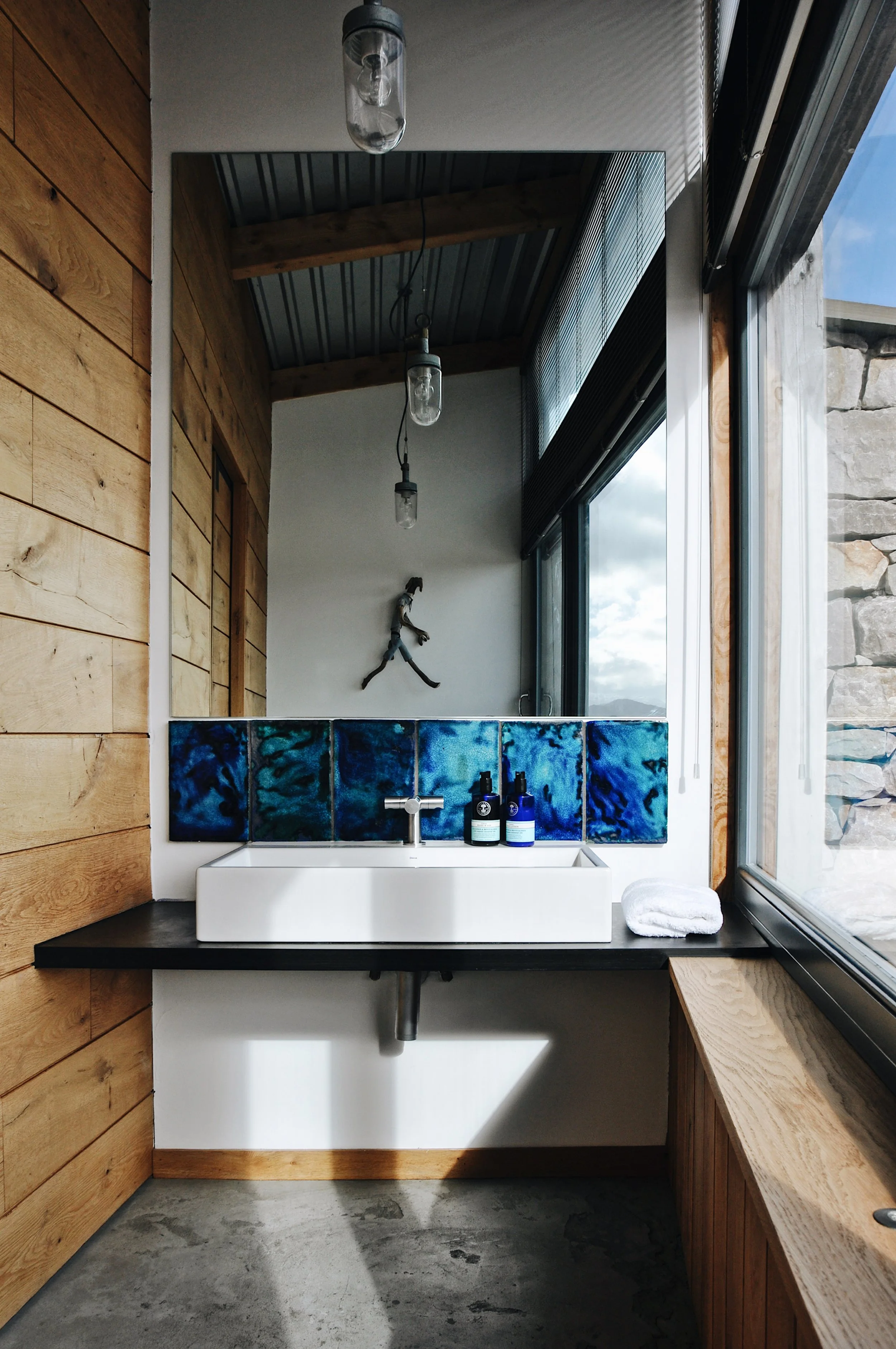 A bathroom with a floating black countertop, a white rectangular vessel sink, and two blue glass bottles. The wall behind the sink has a strip of blue patterned tiles, and there is a large window on the right side. Above the mirror, a metal sculpture of a lizard is mounted on the white wall. The ceiling is made of wooden beams and metal panels, with hanging light fixtures.
