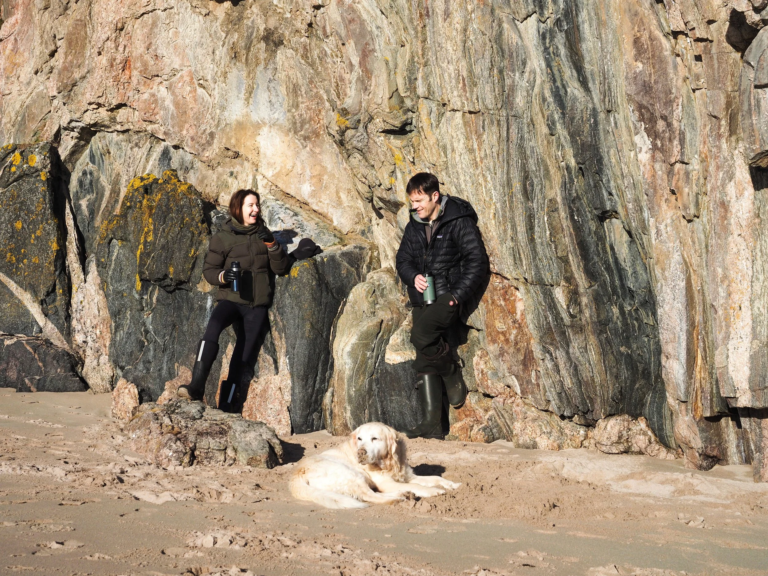 A woman and man in outdoor winter clothing standing on a sandy beach next to large rocks and cliffs, with a golden retriever lying on the sand in front of them. The woman is holding a thermos and a cup, while the man is holding a thermos, and they are smiling at each other.