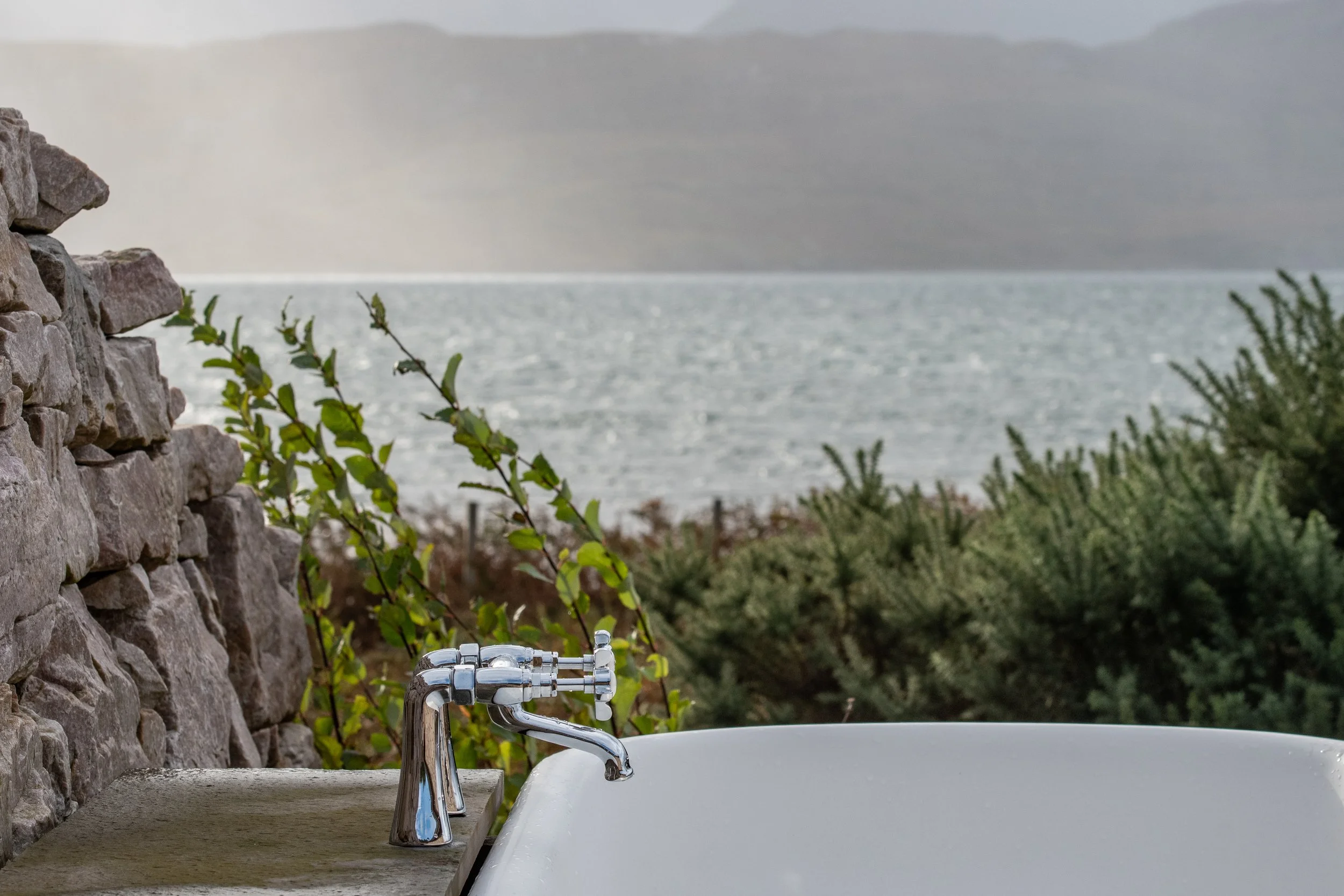 Outdoor bathtub with a chrome faucet near a stone wall, overlooking a body of water with mountains in the distance, and greenery in the foreground.