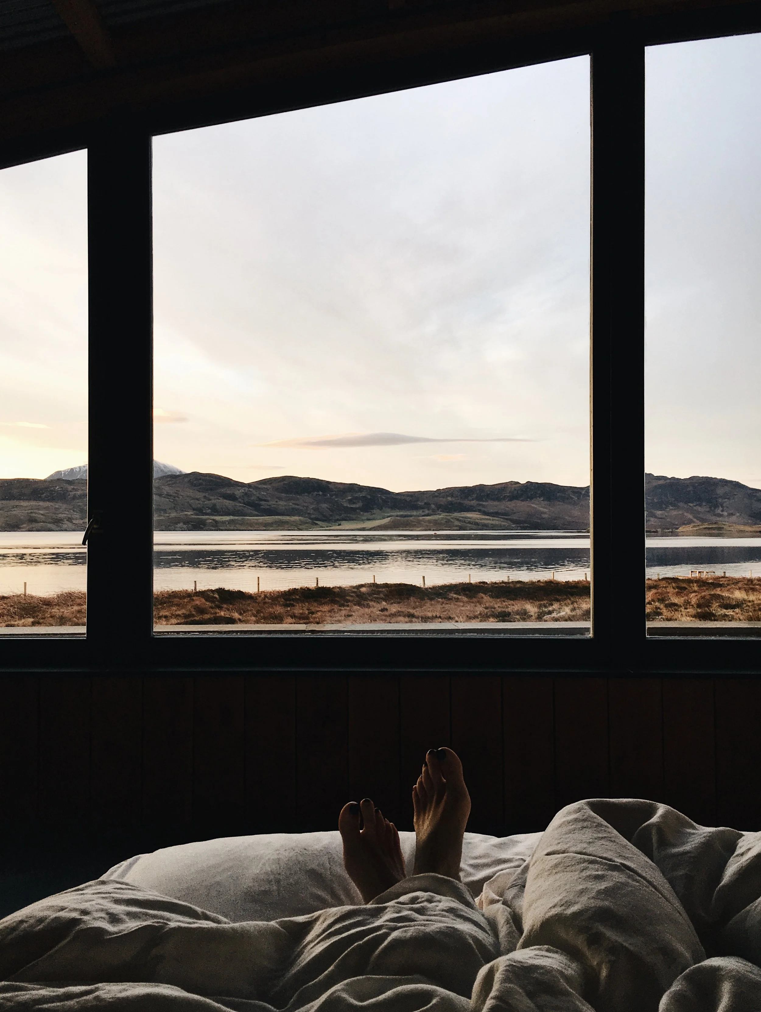 Person lying in bed, looking out a large window at a calm lake and distant mountains on a cloudy day.