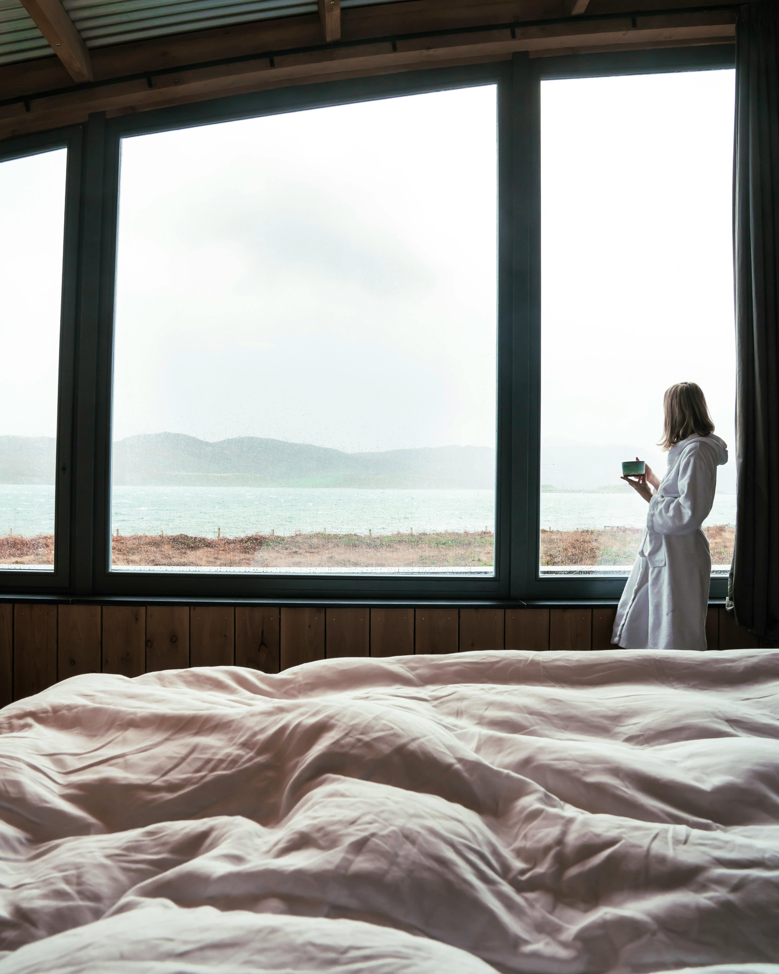 A woman in a white bathrobe standing by a large window, holding a cup, overlooking a body of water and distant hills, with a bedding-covered bed in the foreground.
