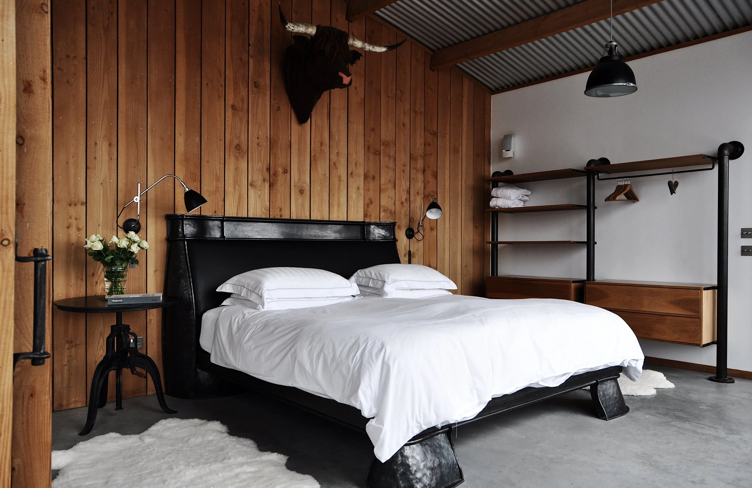 A modern bedroom with a black bed, white bedding, and wooden accents. A mounted animal head decorates the wood-paneled wall, and there are wall-mounted lamps, a small side table with a vase of white roses, and open shelving with towels.