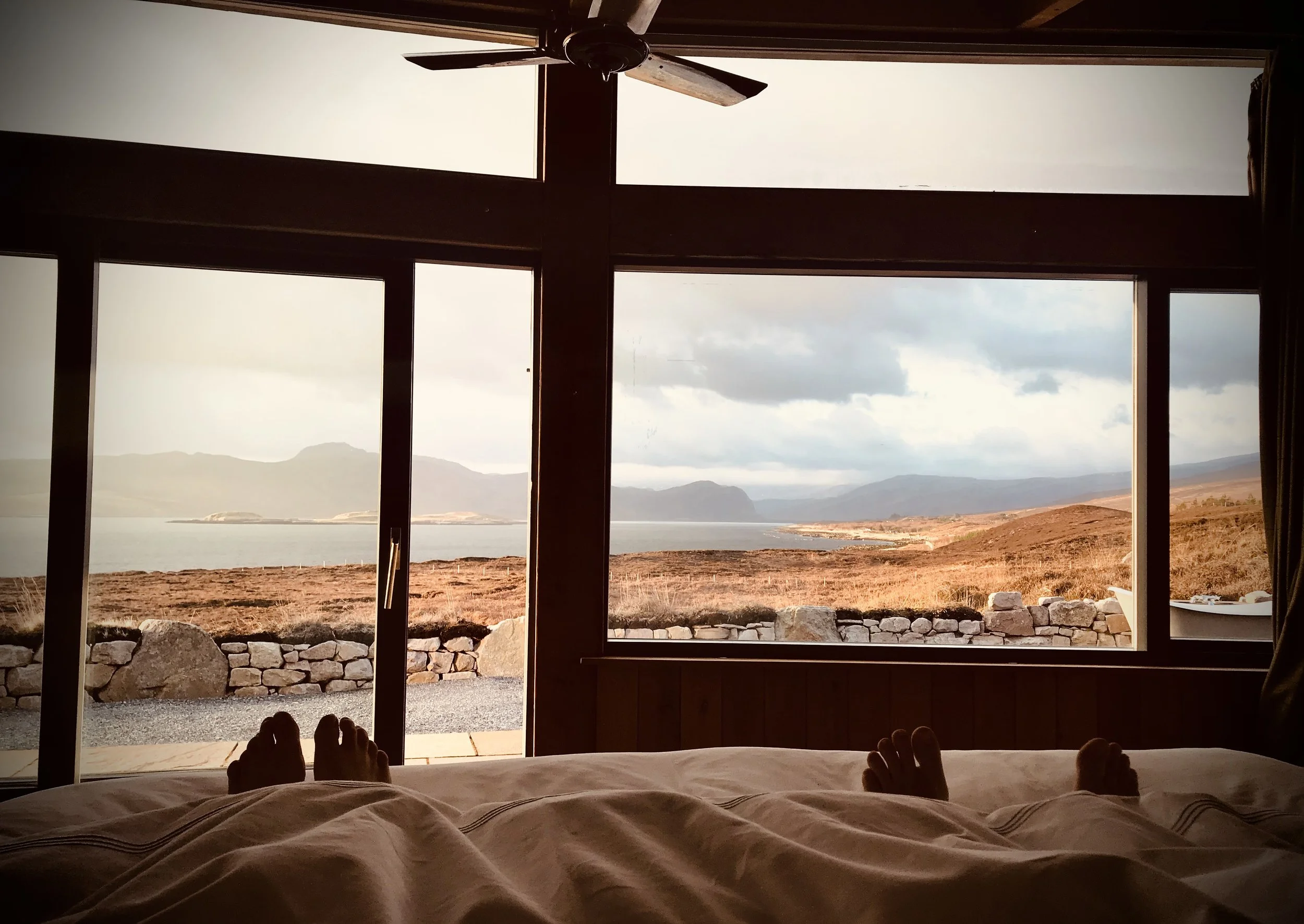 View from a bed showing feet, looking out a large window at a lake, mountains, and cloudy sky during sunset.