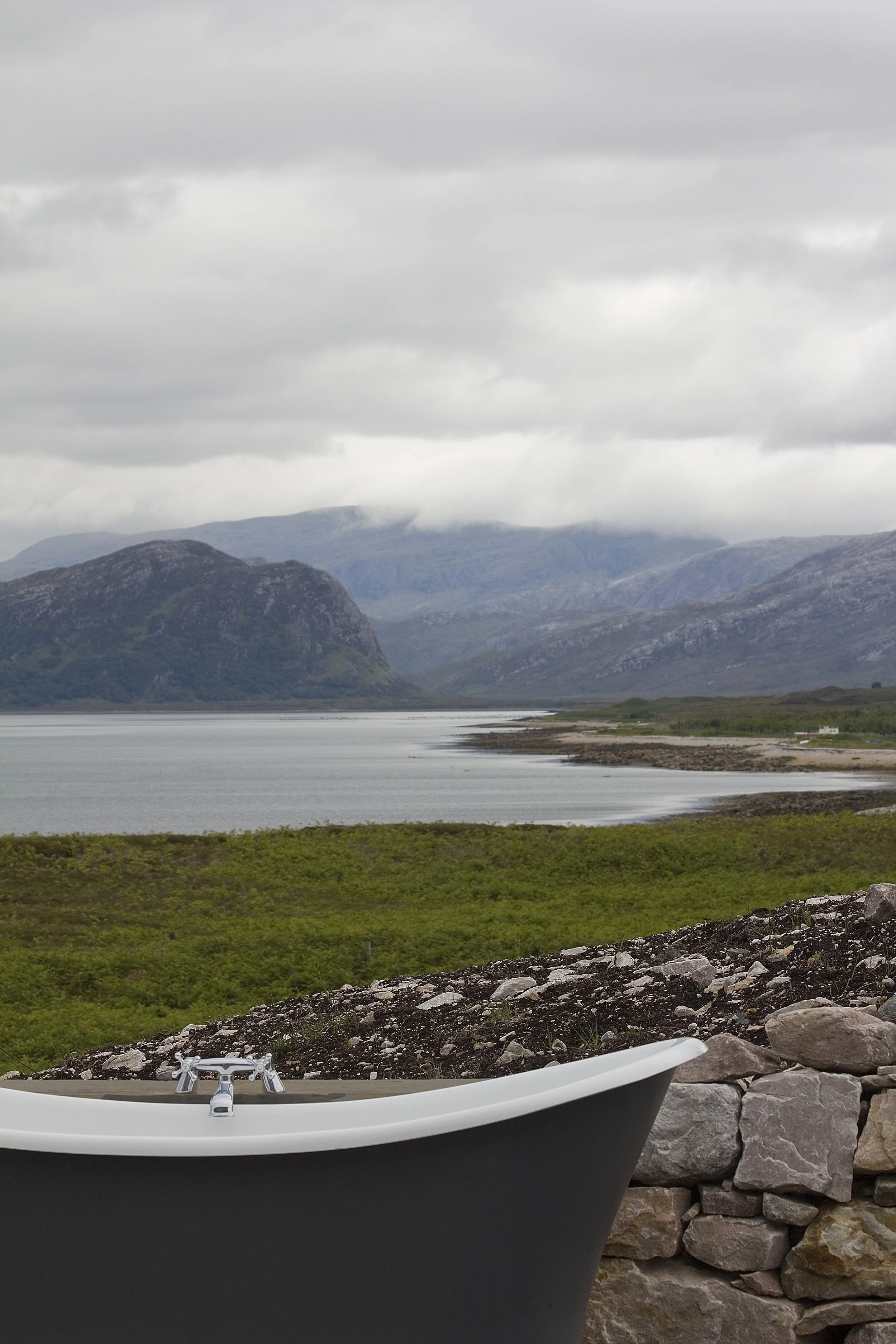 An outdoor scene featuring a white bathtub with silver fixtures in the foreground, a stone wall, and a scenic landscape with a lake, green fields, and mountains under a cloudy sky in the background.