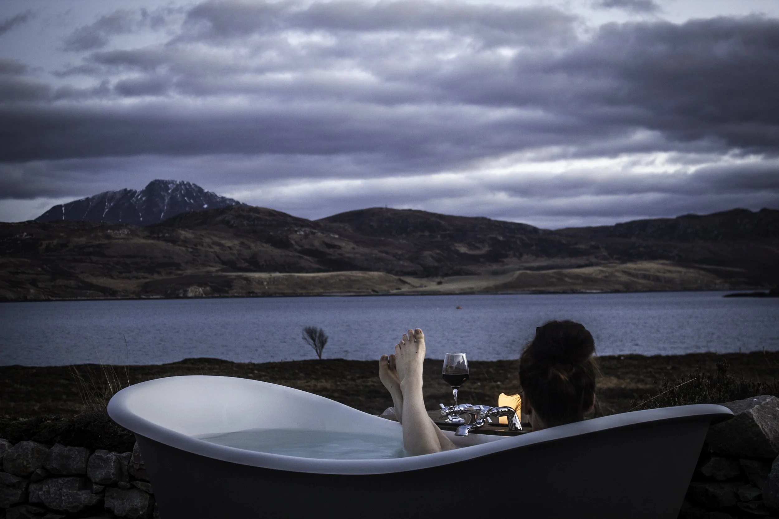A woman relaxing in a bathtub outdoors by a lake during dusk, with mountains in the background and a glass of red wine nearby.