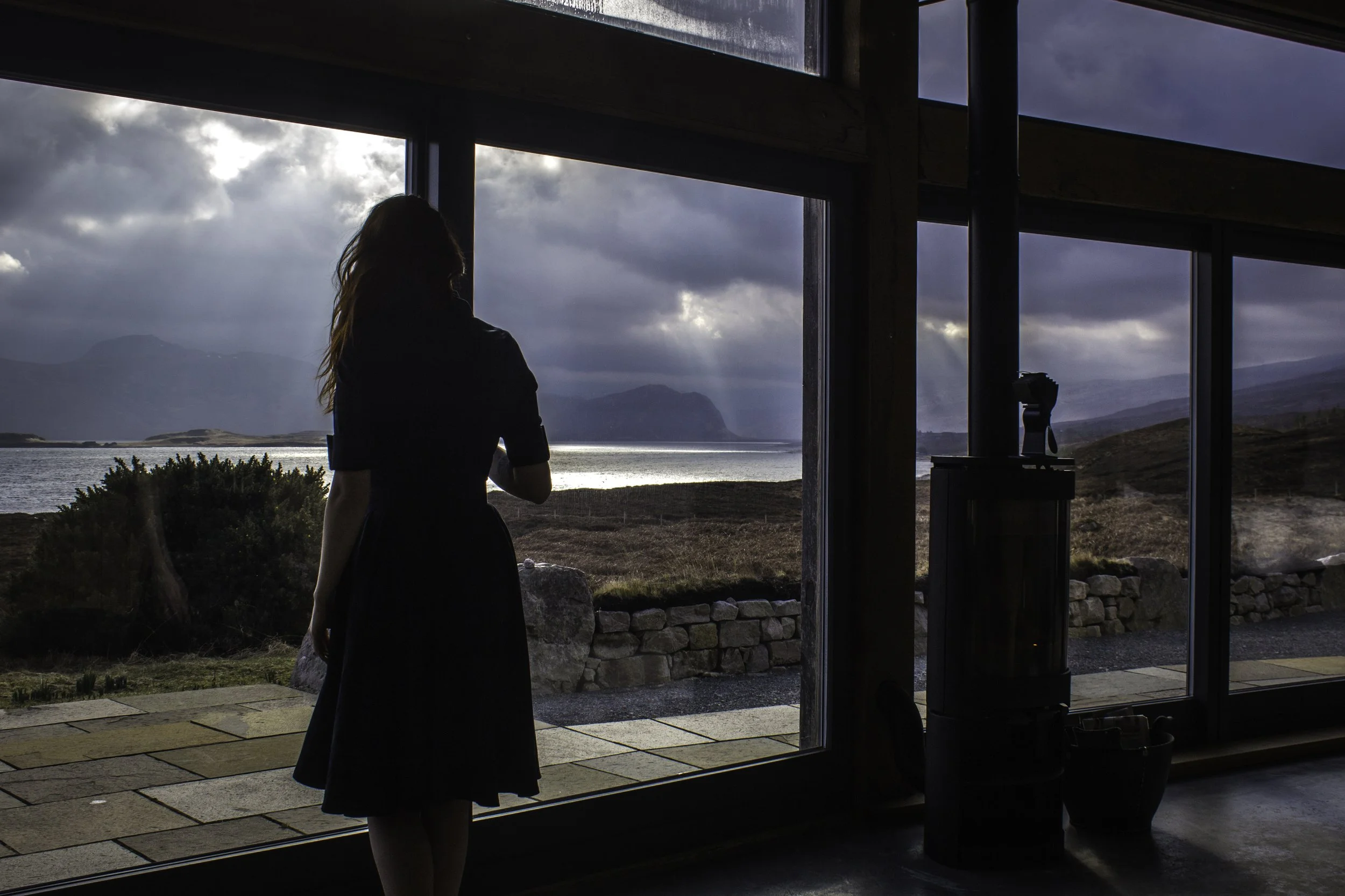 A woman standing inside near large glass windows, looking out at a scenic view of a lake, mountains, and cloudy sky.