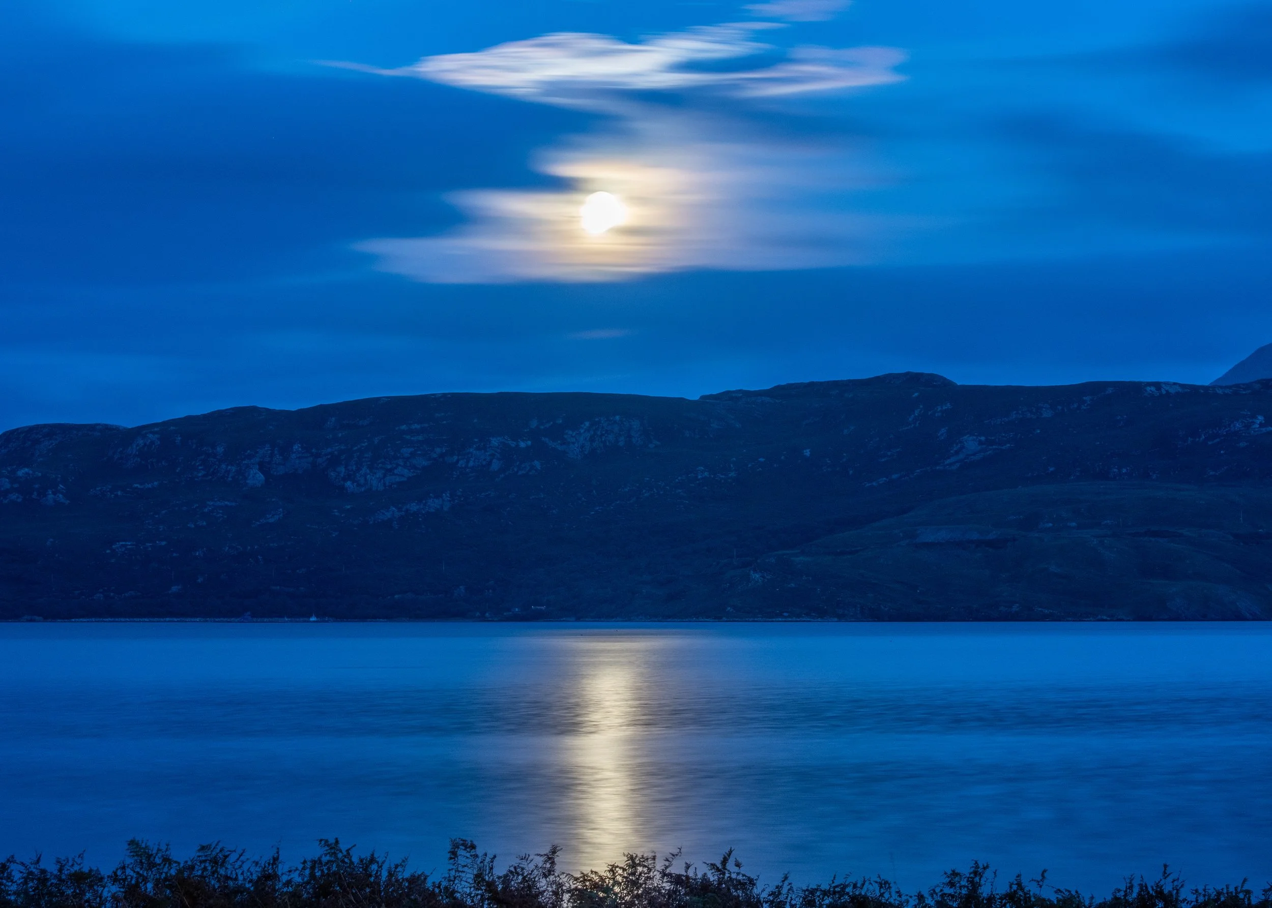 Nighttime scene of a full moon reflecting on a calm lake, with hills in the background and clouds in the sky.