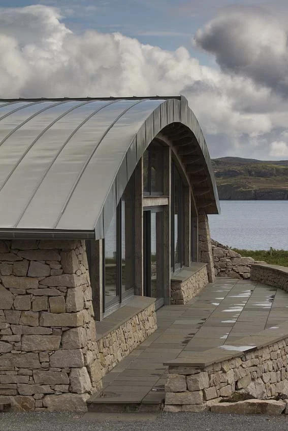 A stone building with large glass windows and a rounded metal roof, situated outdoors near water and hills under a cloudy sky.