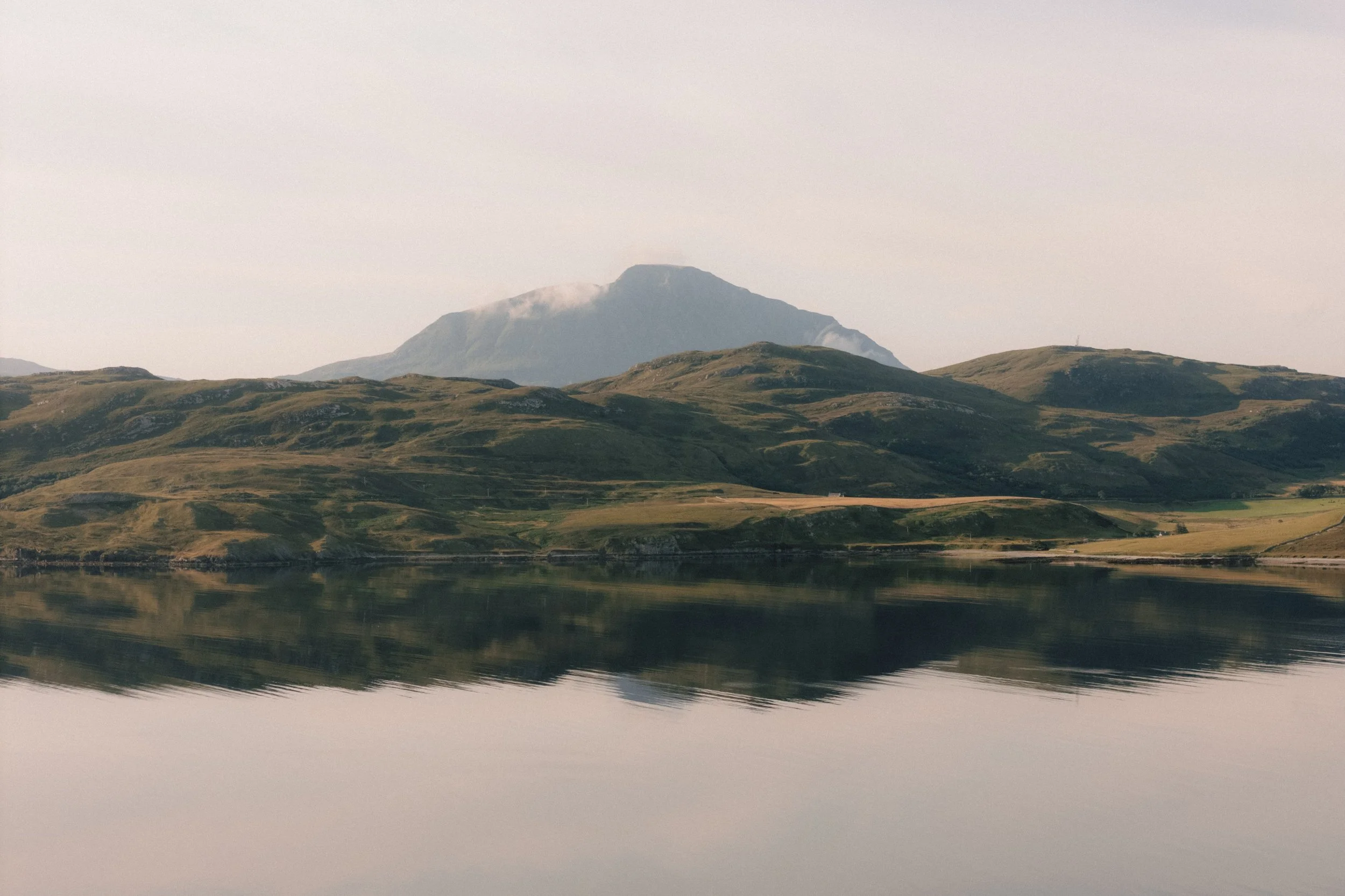 Scenic view of rolling green hills reflected in a calm lake with a mountain in the background under a light sky.