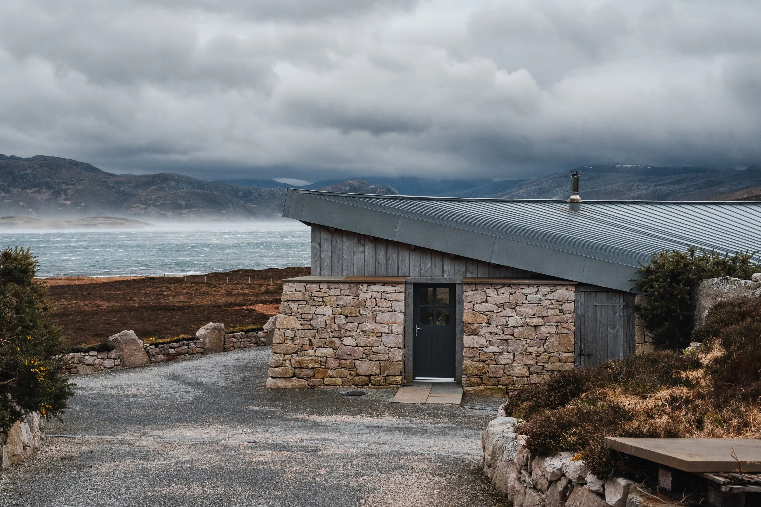 A stone and wood cabin with a metal roof on a gravel driveway, overlooking a lake and mountain landscape under cloudy skies.