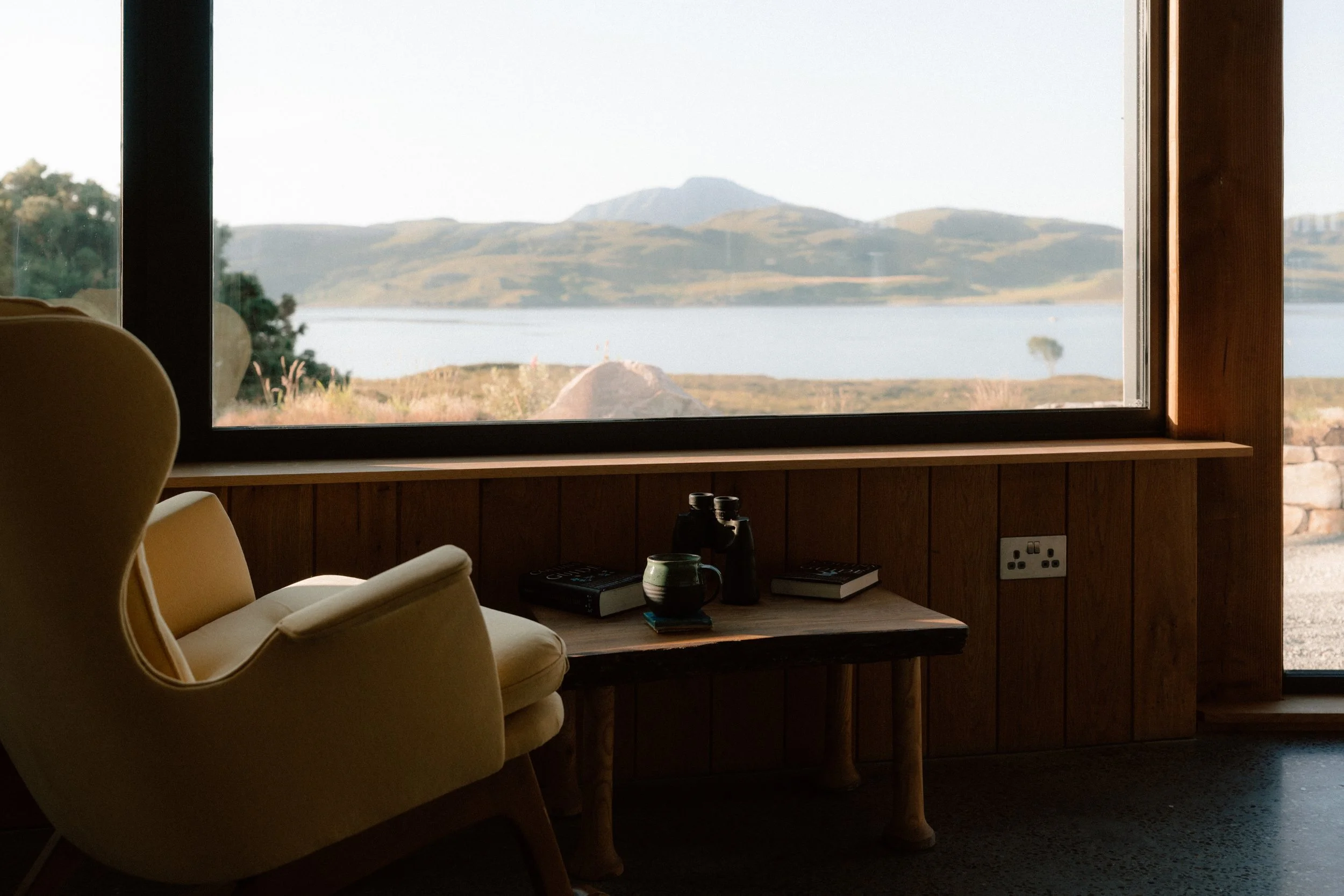 Interior of a room with a large window overlooking a lake and mountains, a cream-colored armchair, a wooden table with books, binoculars, a cup, and a remote control, and an electrical outlet on the wooden wall.