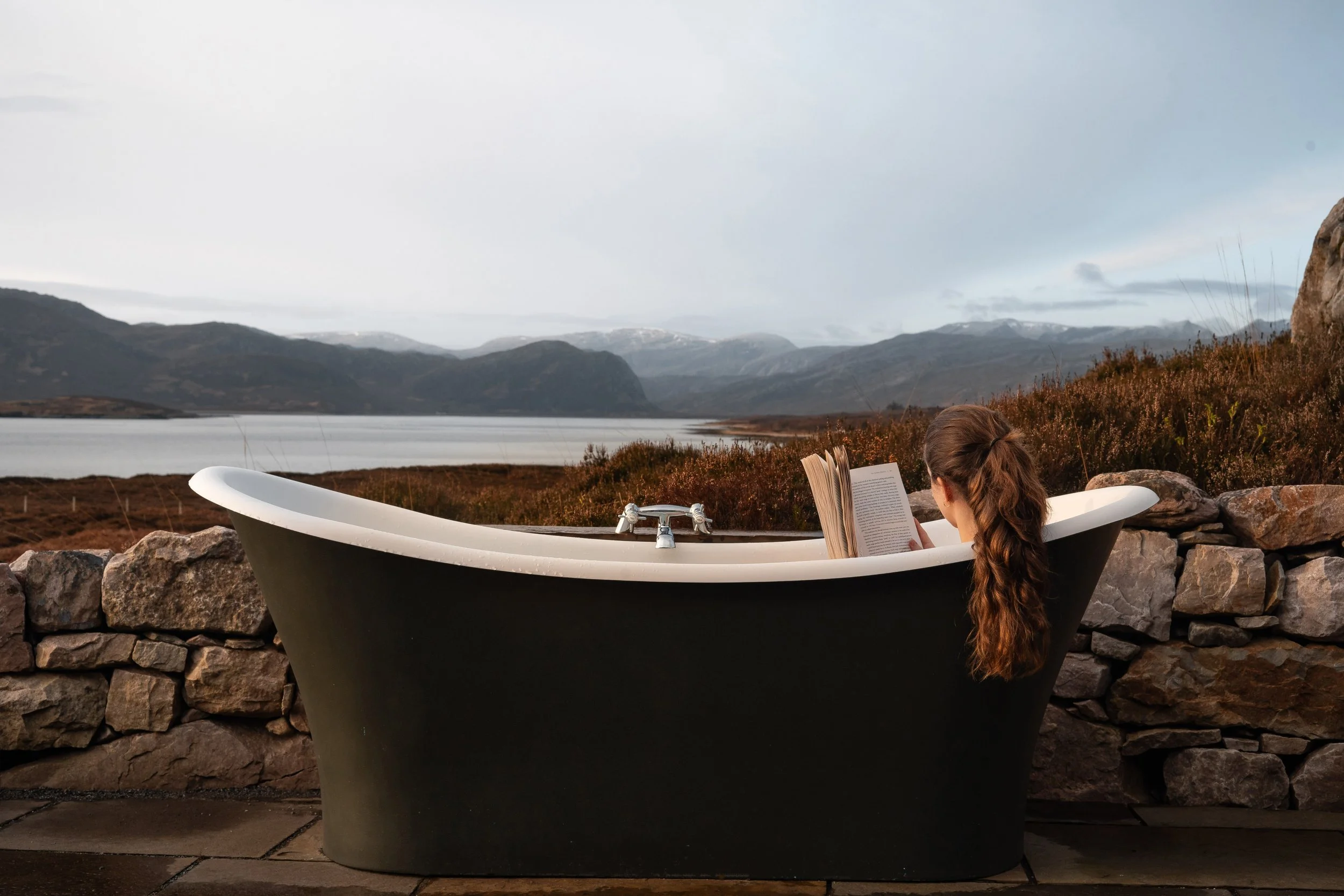 A woman with long brown hair reading a book in a black and white bathtub outdoors near a stone wall, overlooking a lake and mountains under a cloudy sky.