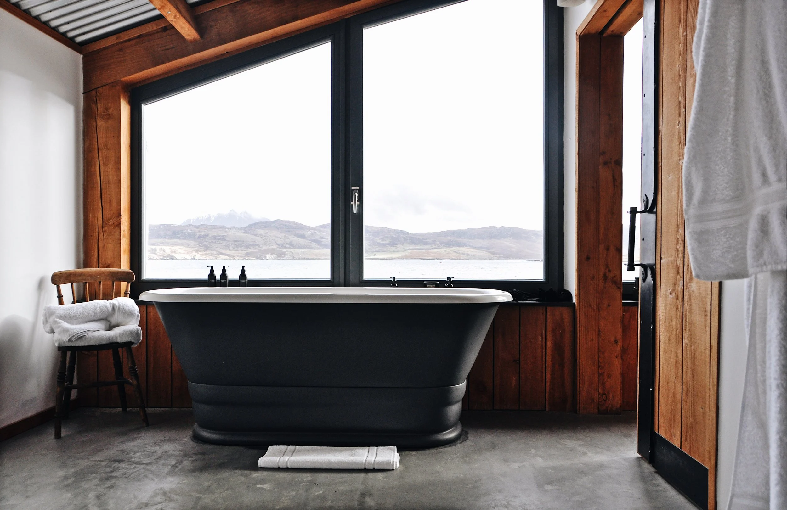 A rustic bathroom with a black bathtub placed in front of large windows overlooking mountains and a lake, with a wooden chair to the left and towels hanging on the door.