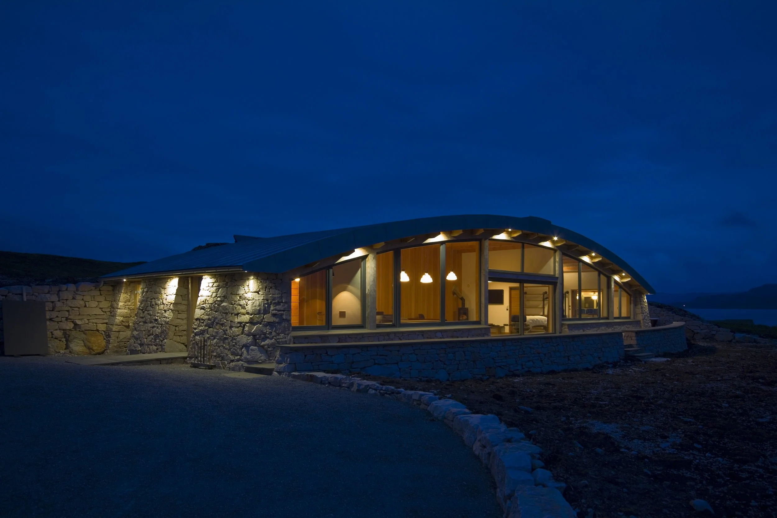 A modern house with large glass windows and a curved roof at dusk, built with stone walls and warm interior lighting.