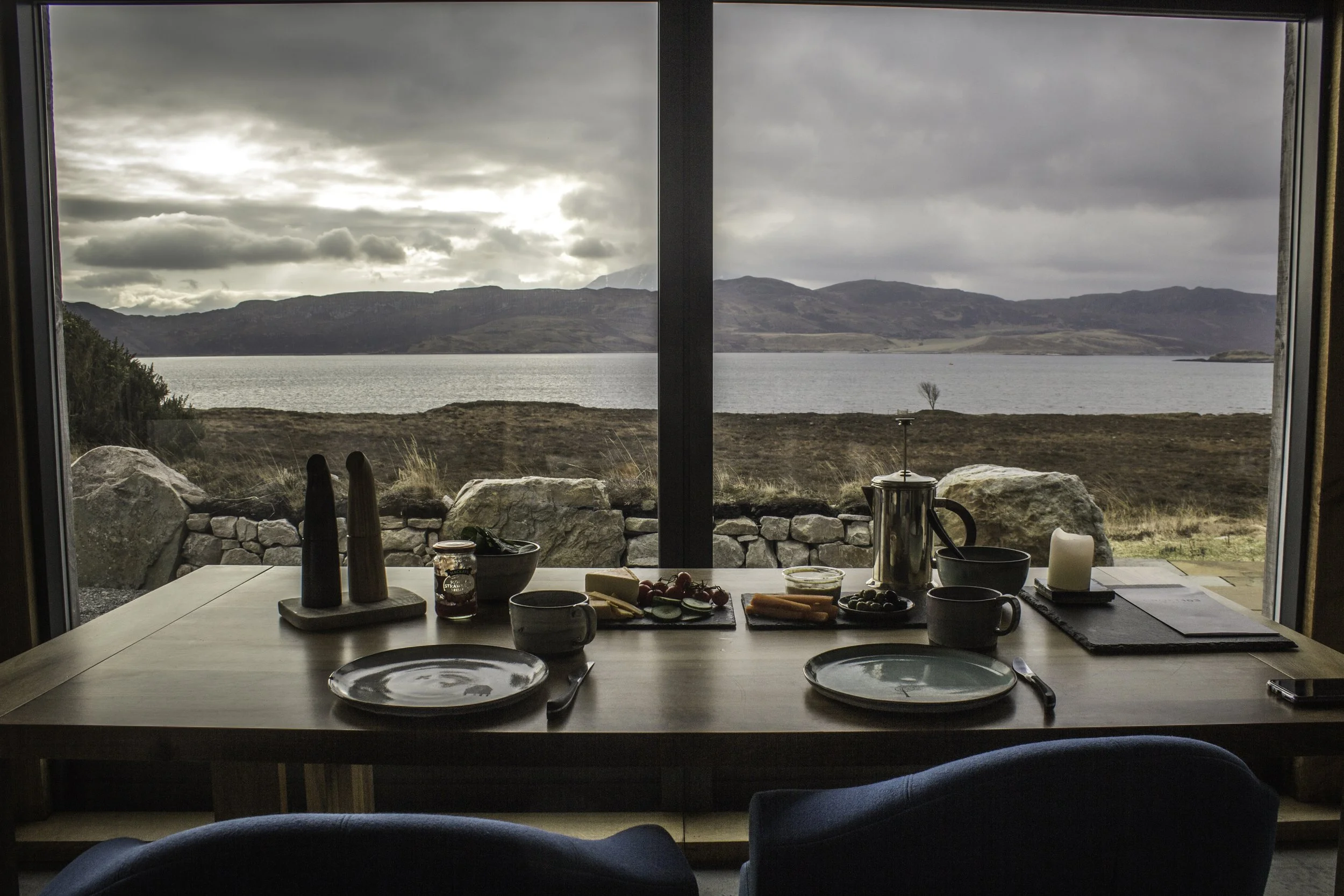 Breakfast table set with plates, cups, and food, overlooking a lake and mountains through large window.