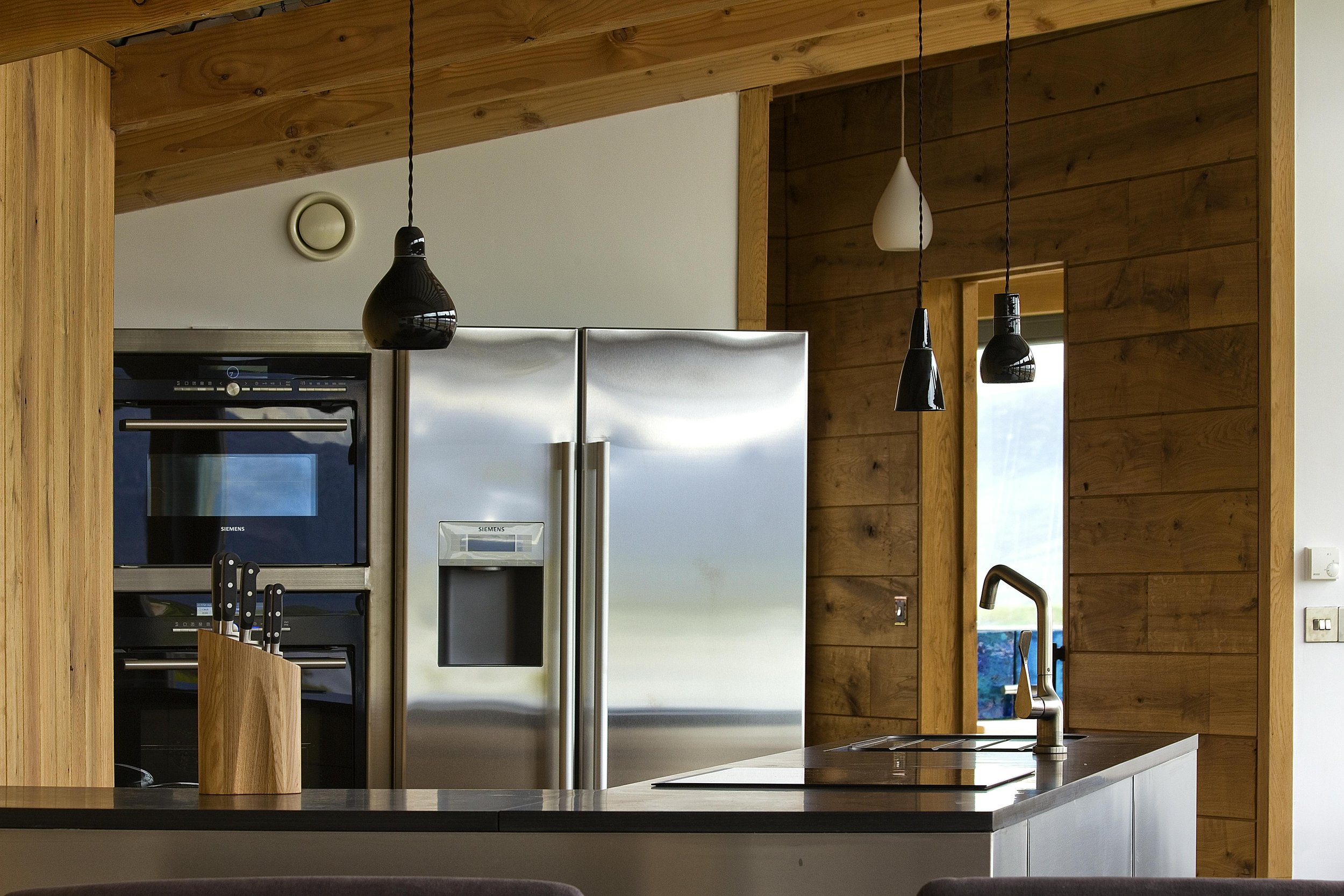 Modern kitchen with stainless steel refrigerator, wooden walls, black pendant lights, and a kitchen island with a sink.