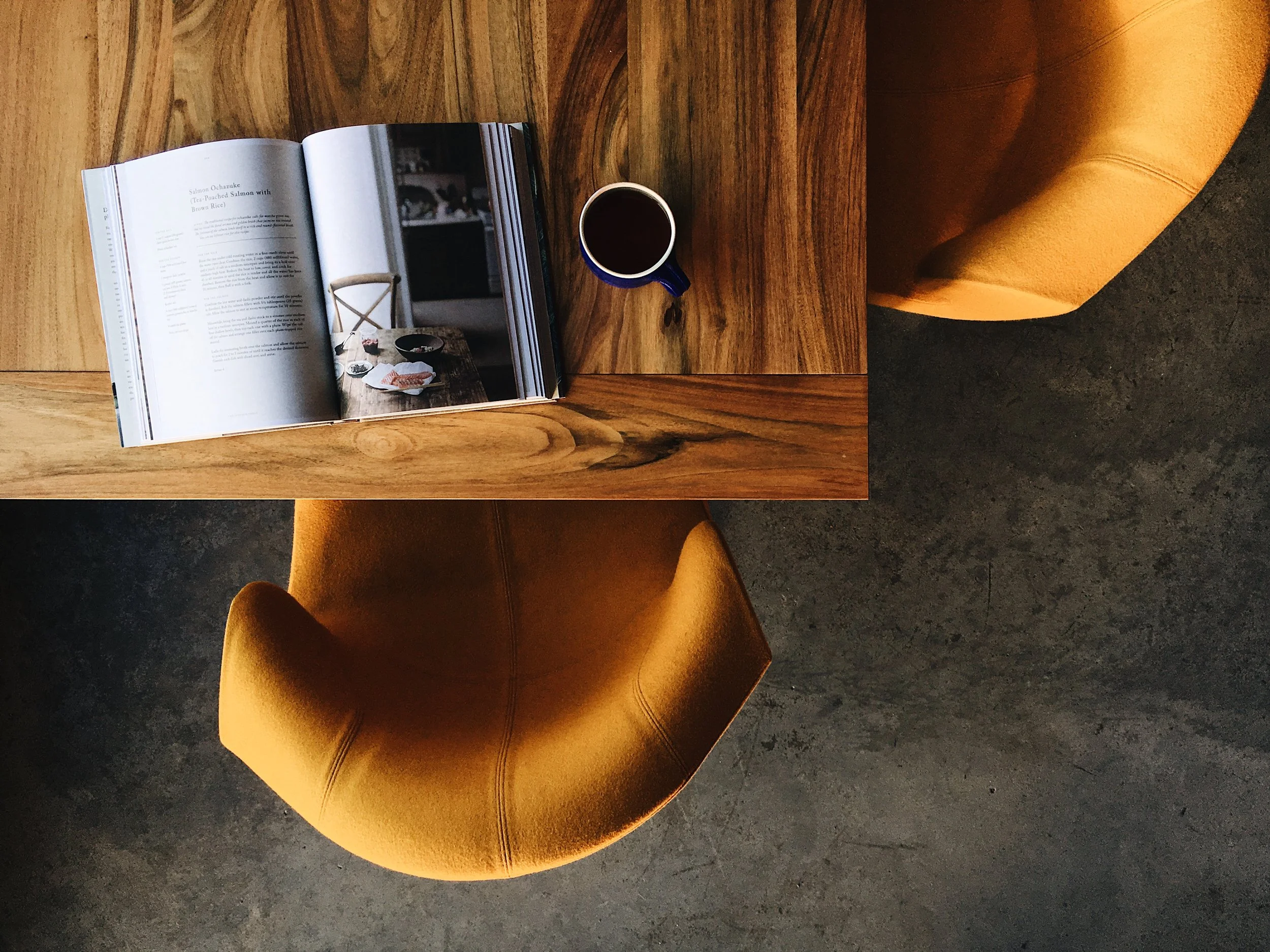 A top-down view of a wooden table with an open magazine, a blue cup of coffee, and yellow upholstered chairs.