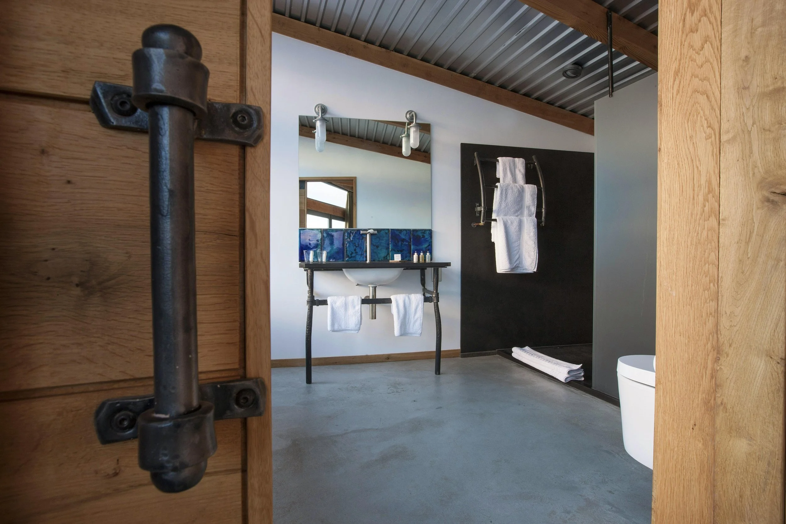 View of a modern bathroom seen through a wooden door frame with a metal latch. The bathroom features a black countertop sink with blue tile backsplash, a large mirror, white towels hanging underneath, white towels on the floor, a black wall with a towel rack holding two white towels, a roll of towels, a white toilet, and a metal ceiling with wooden beams.