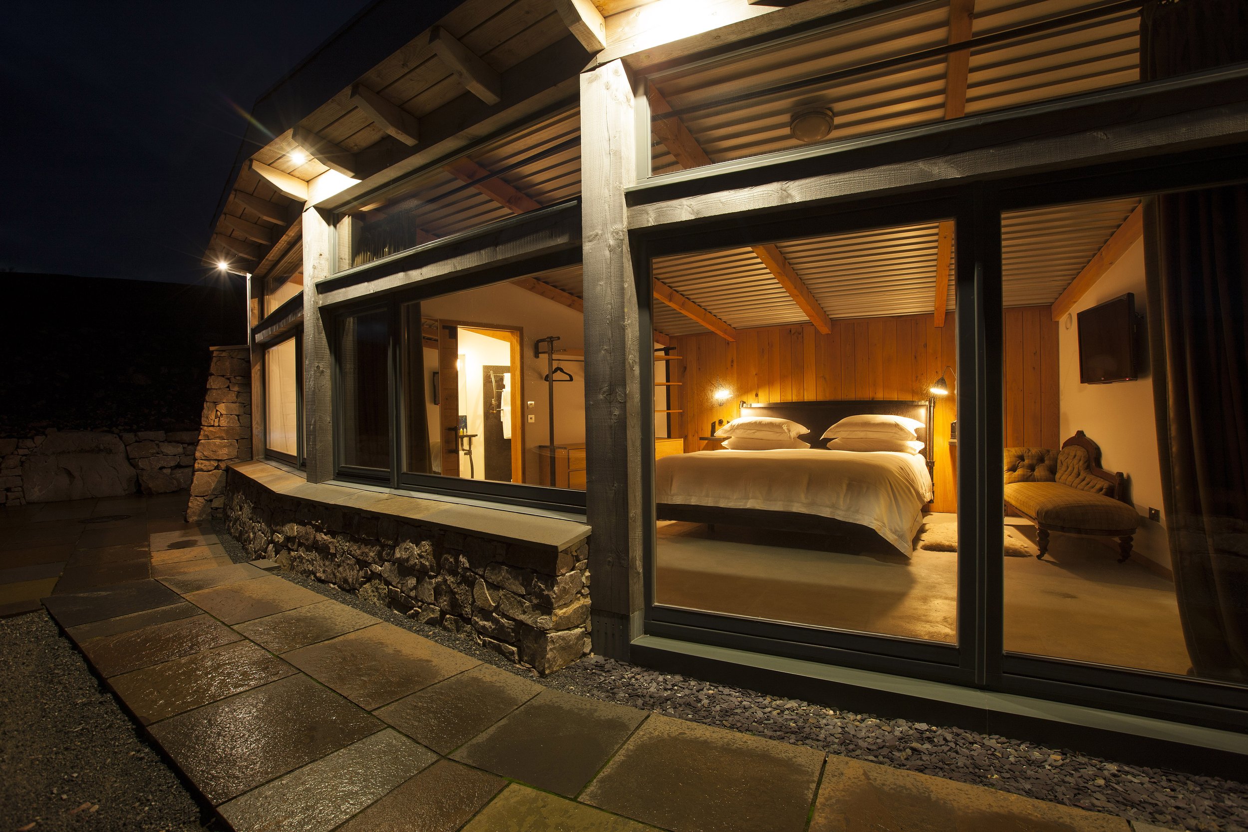 View of a cozy, well-lit bedroom in a rustic house at night, with large glass windows showing the interior with a bed, wooden walls, and furniture.