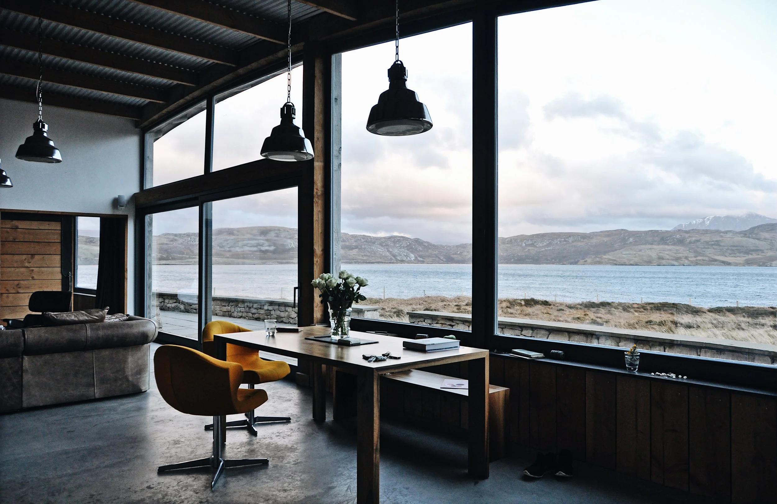Interior of a modern room with large windows showcasing a scenic view of a lake and mountains, furnished with a wooden table, yellow chairs, a gray sofa, and hanging black lamps.