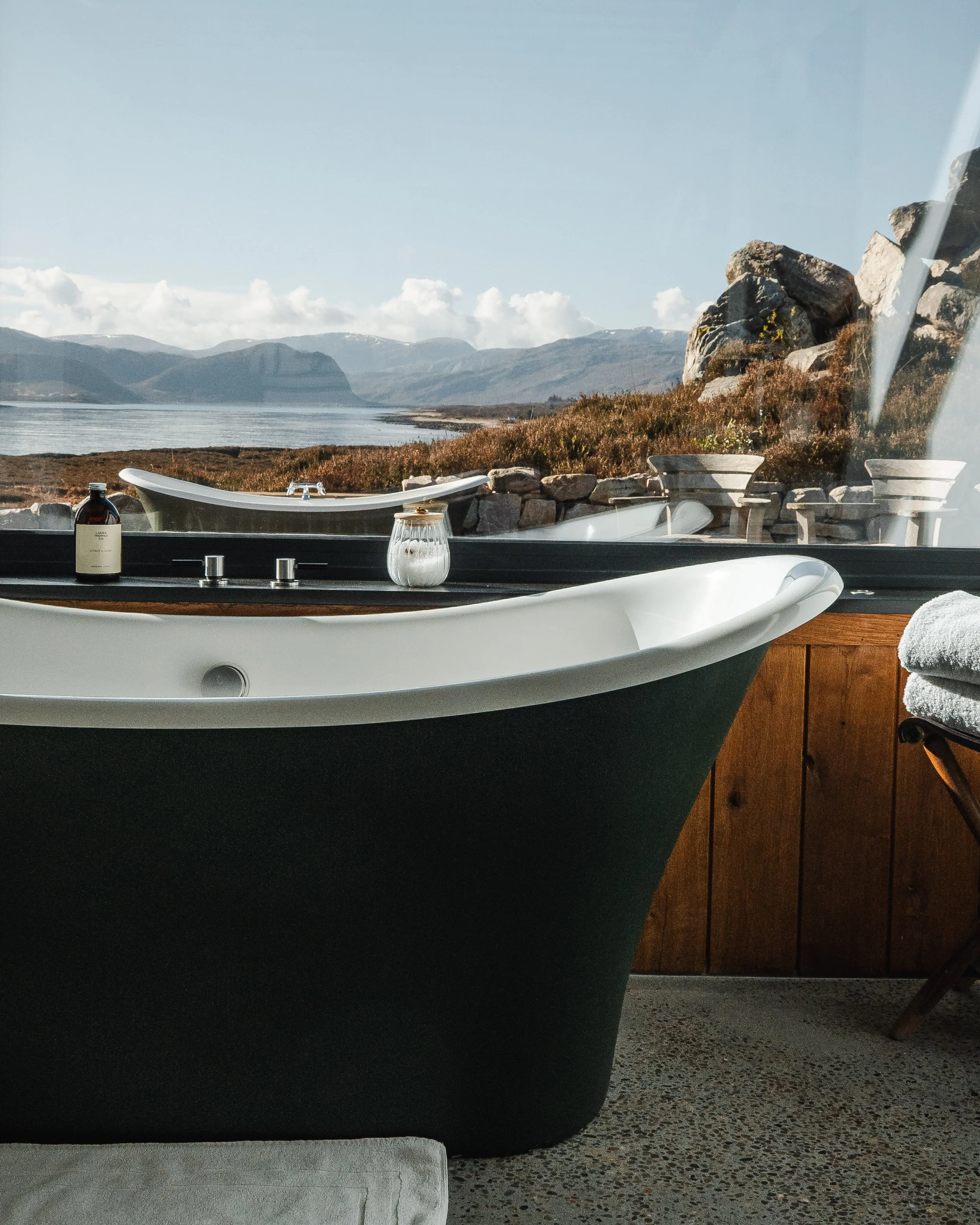 A bathtub with a mountain and lake view outside a window, with a bottle and candle on the tub's edge, and a wooden wall behind.
